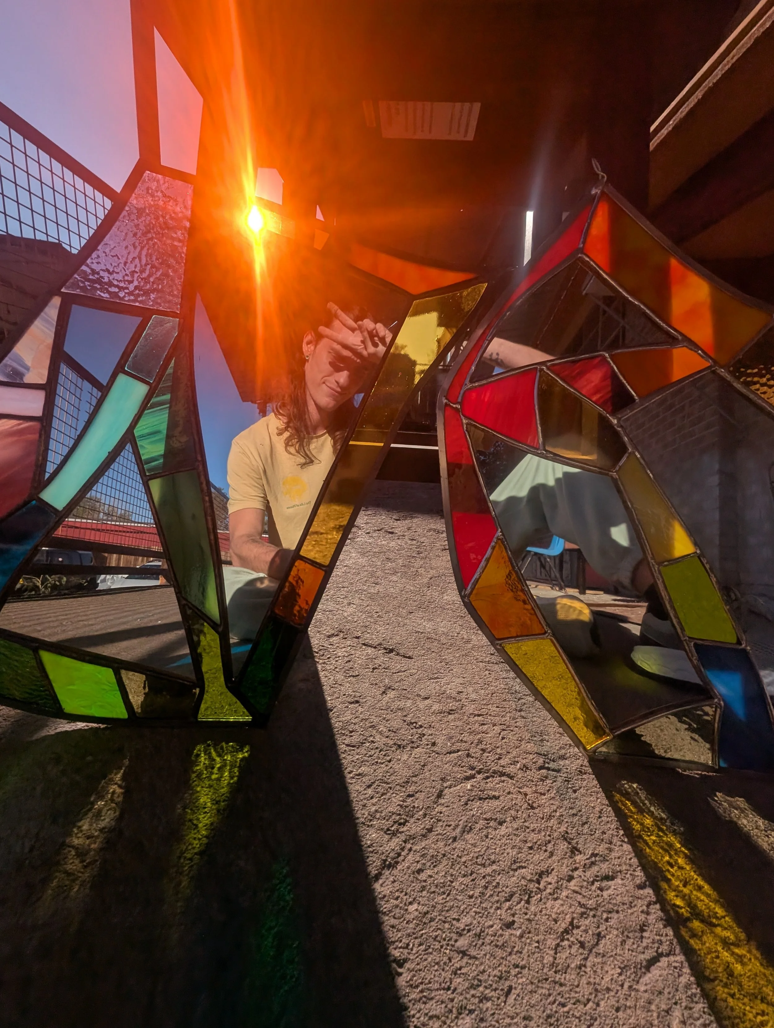 Colorful stained glass installation with a person in a yellow shirt behind it, sunlight creates reflections and shadows on the ground.