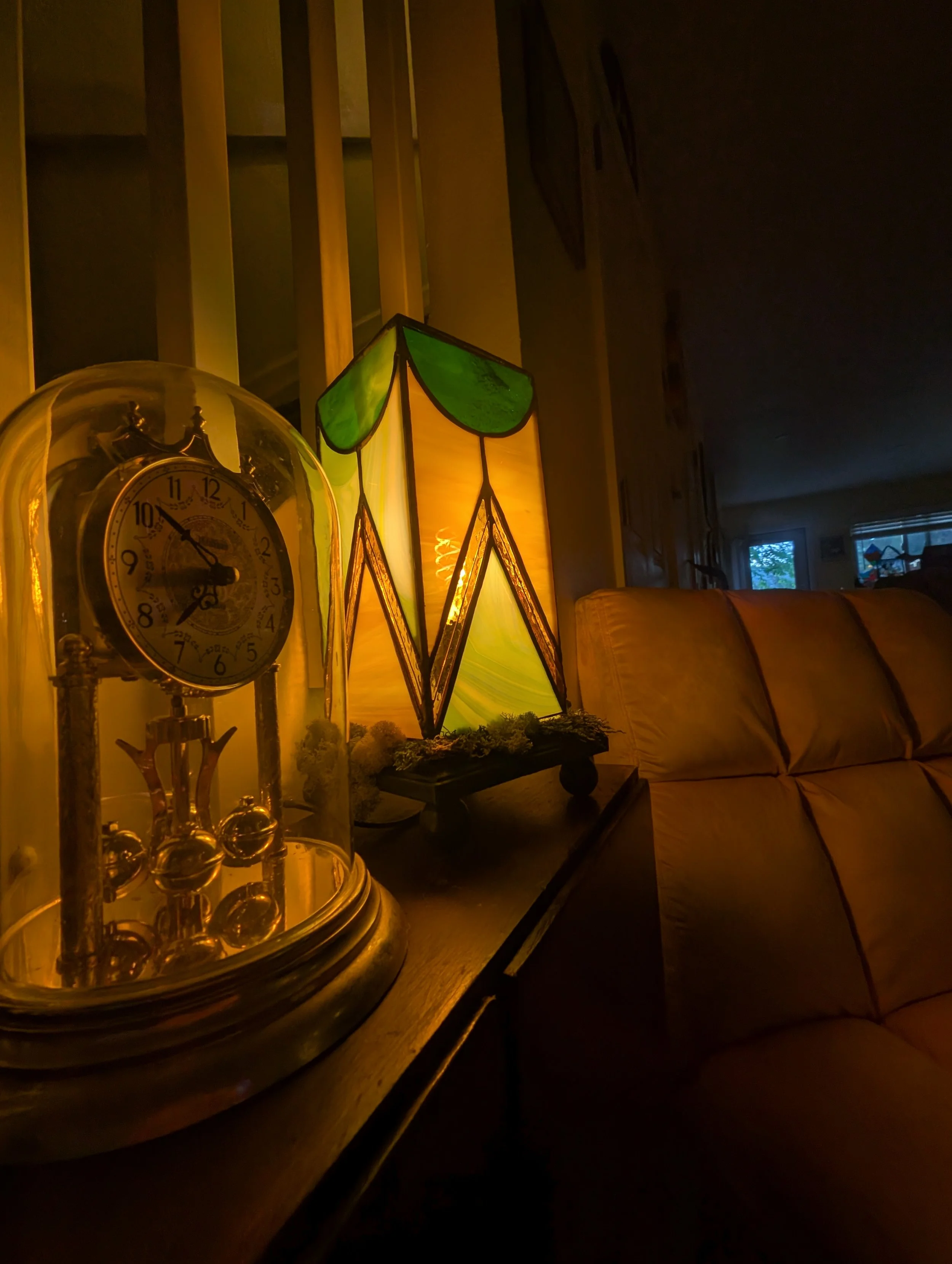A dimly lit living room side table with a vintage clock under a glass dome, a stained glass lamp, and a sofa with beige cushions visible in the background.