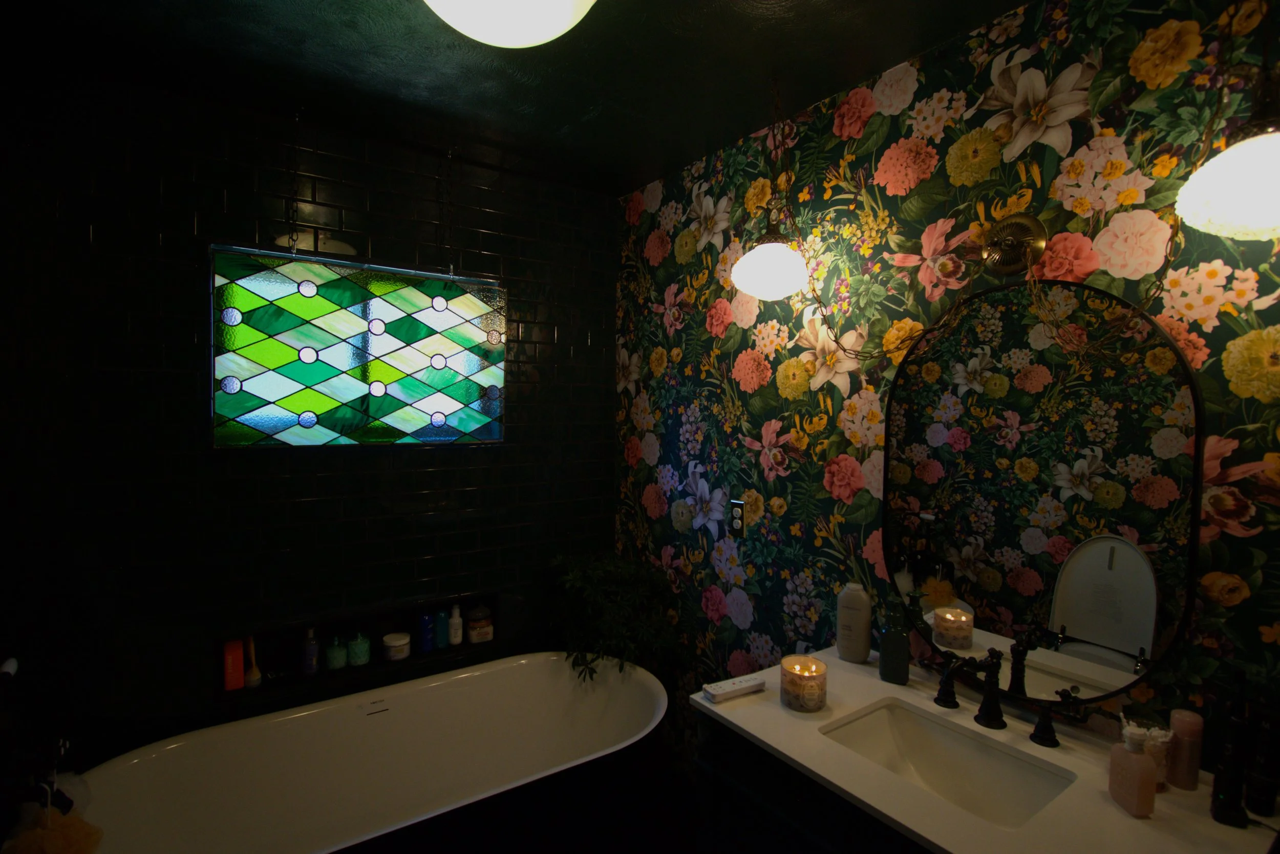 Bathroom with floral wallpaper, oval mirror, lit by two wall sconces, black tiles on one wall, stained glass window, bathtub, and candles on the countertop.
