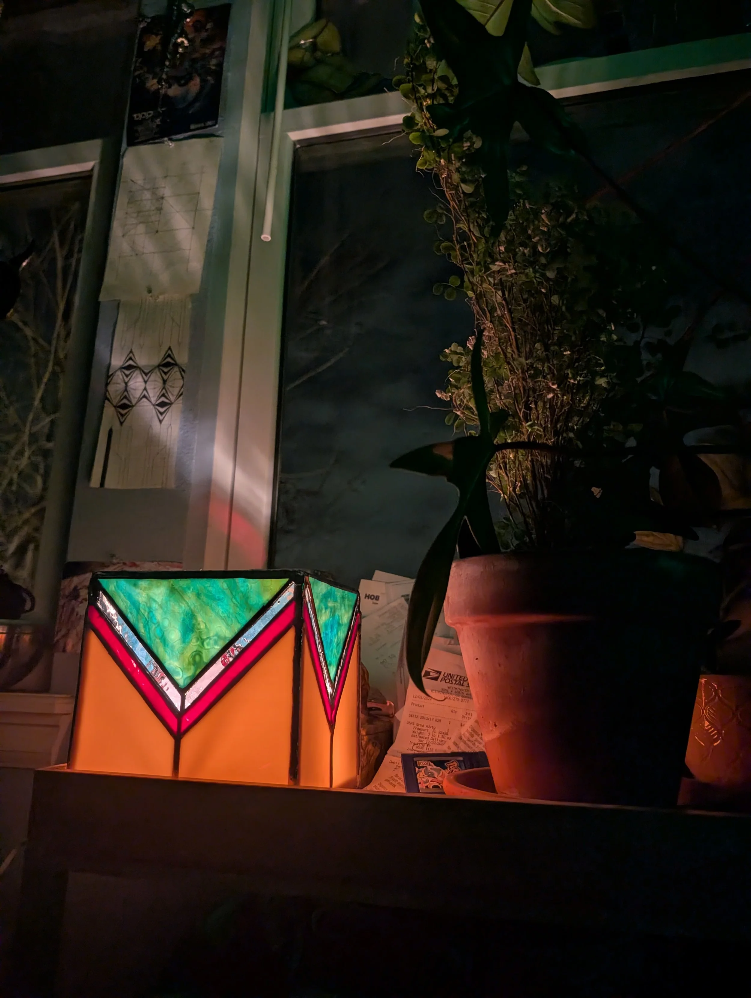 A nighttime scene showing a colorful stained glass-style lamp with geometric patterns, and a potted plant with green leaves, placed on a surface near a window with a dark sky visible outside.