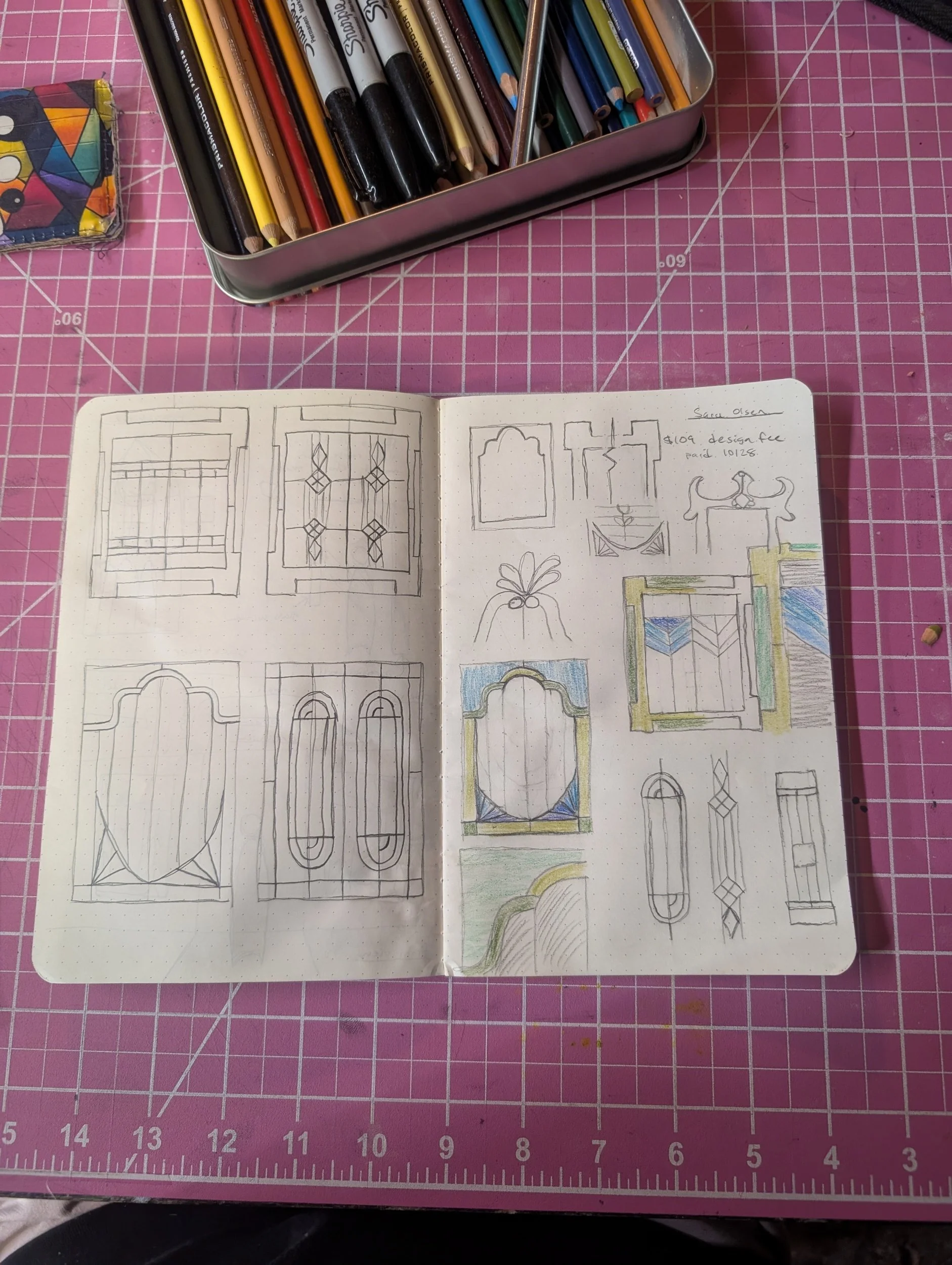 Open sketchbook on pink cutting mat with drawings of window and door designs, some colored with colored pencils, and a box of colored pencils nearby.