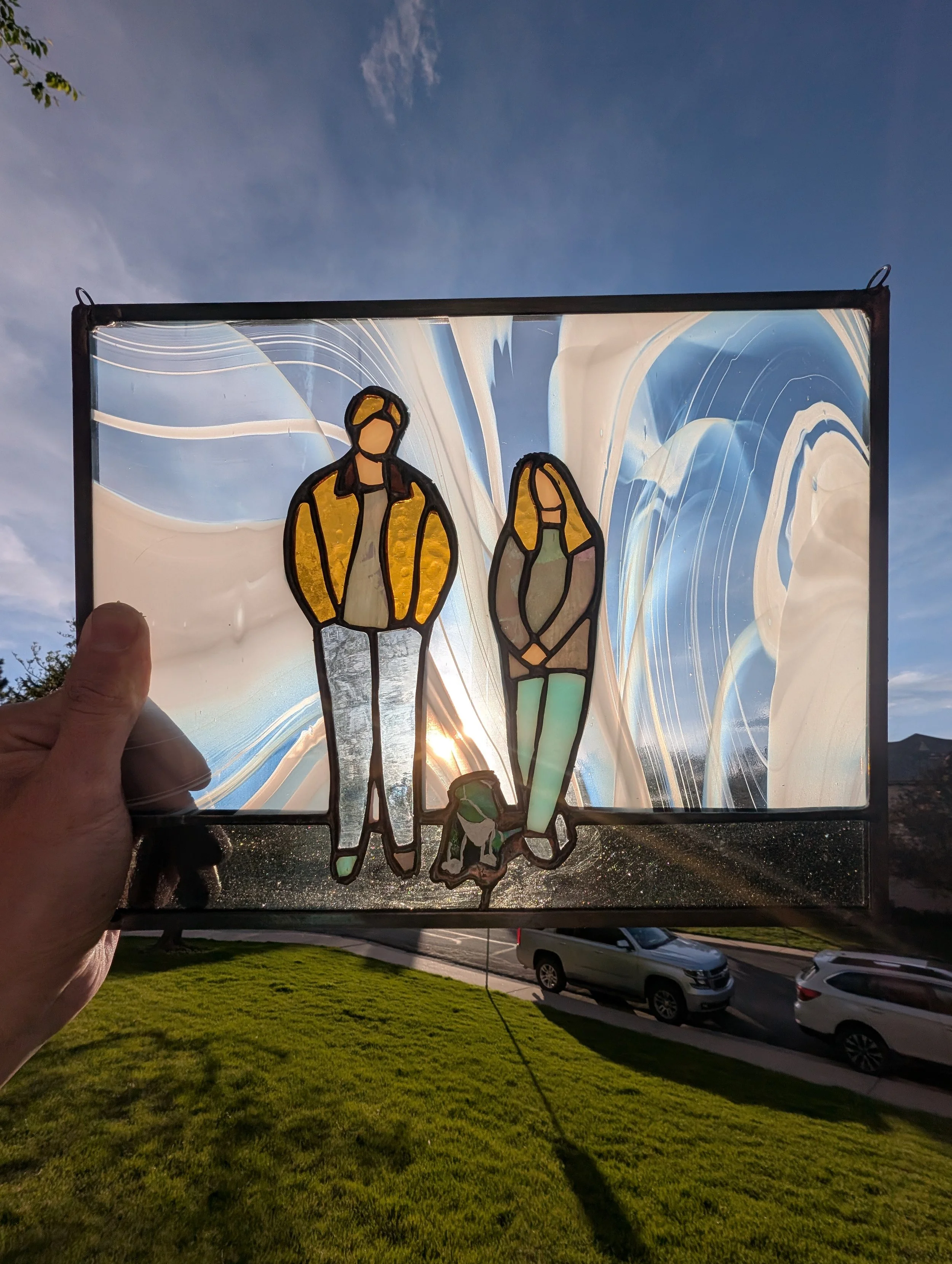 A hand holding a stained glass window with a silhouette of a young man and woman walking together, with cars and a grassy area in the background.