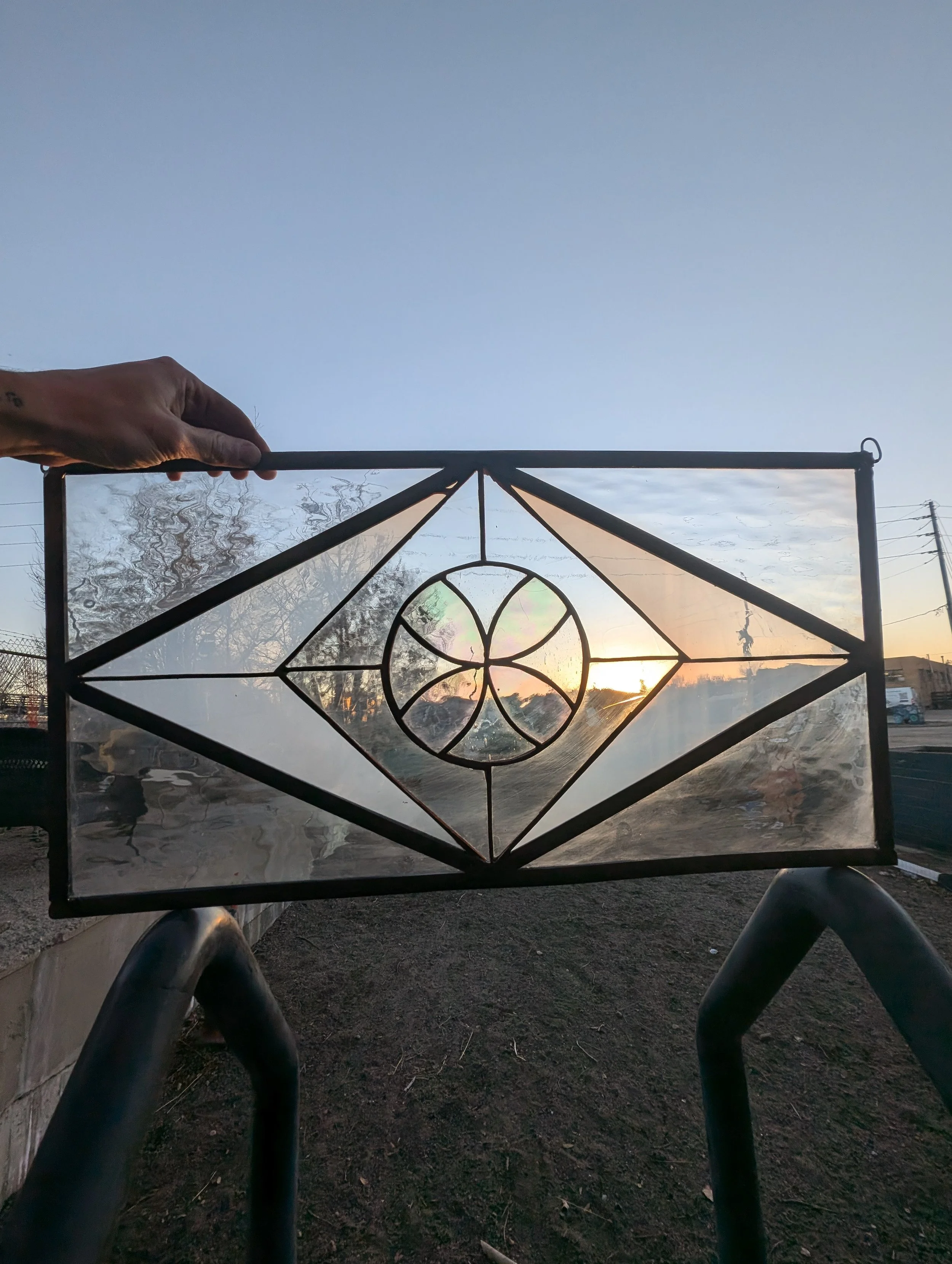 A hand holding a stained glass window with geometric patterns, silhouetted against a sunset sky.