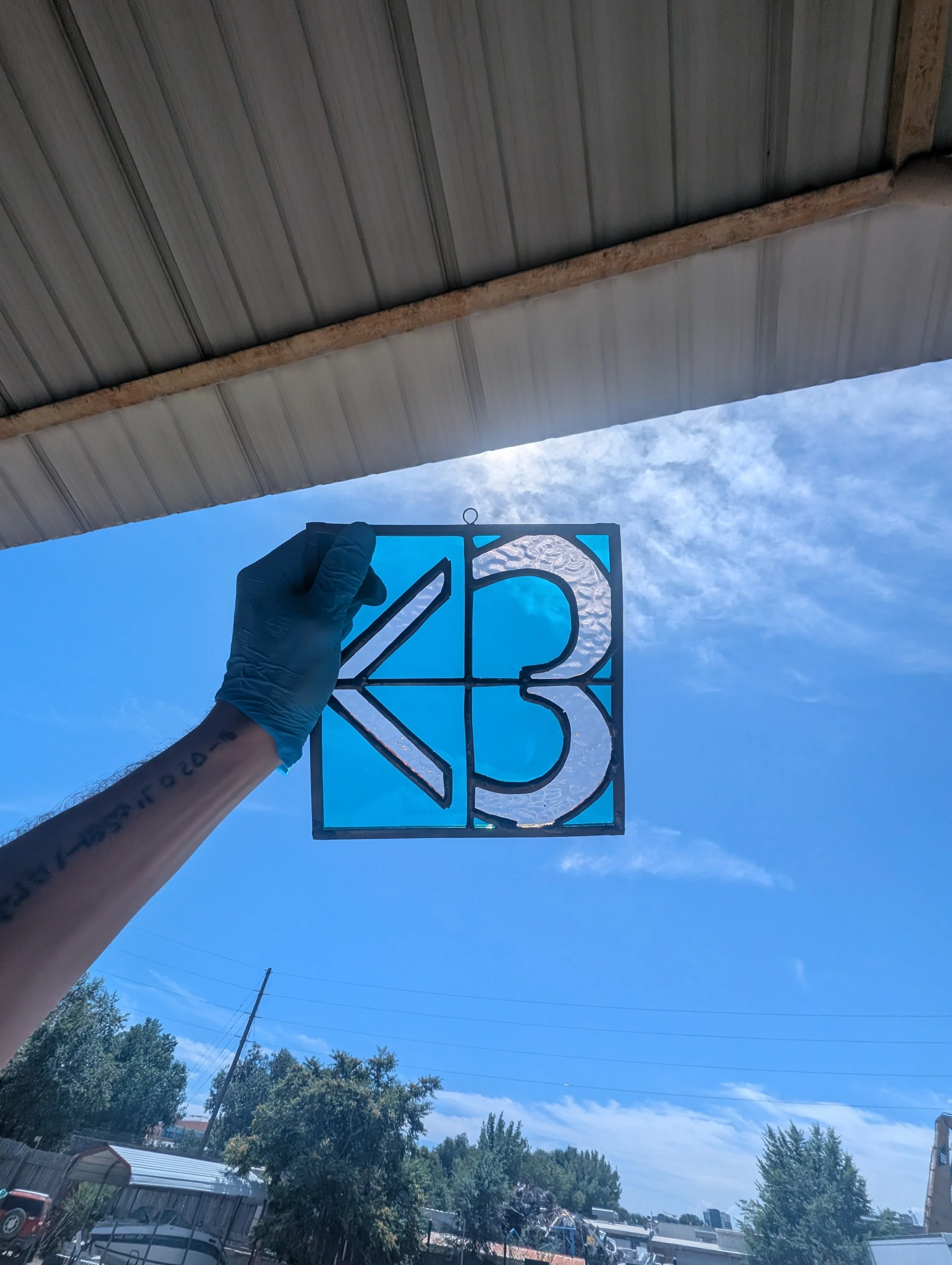 Person holding a stained glass piece with the letters 'K' and the number '3' against a blue sky background.