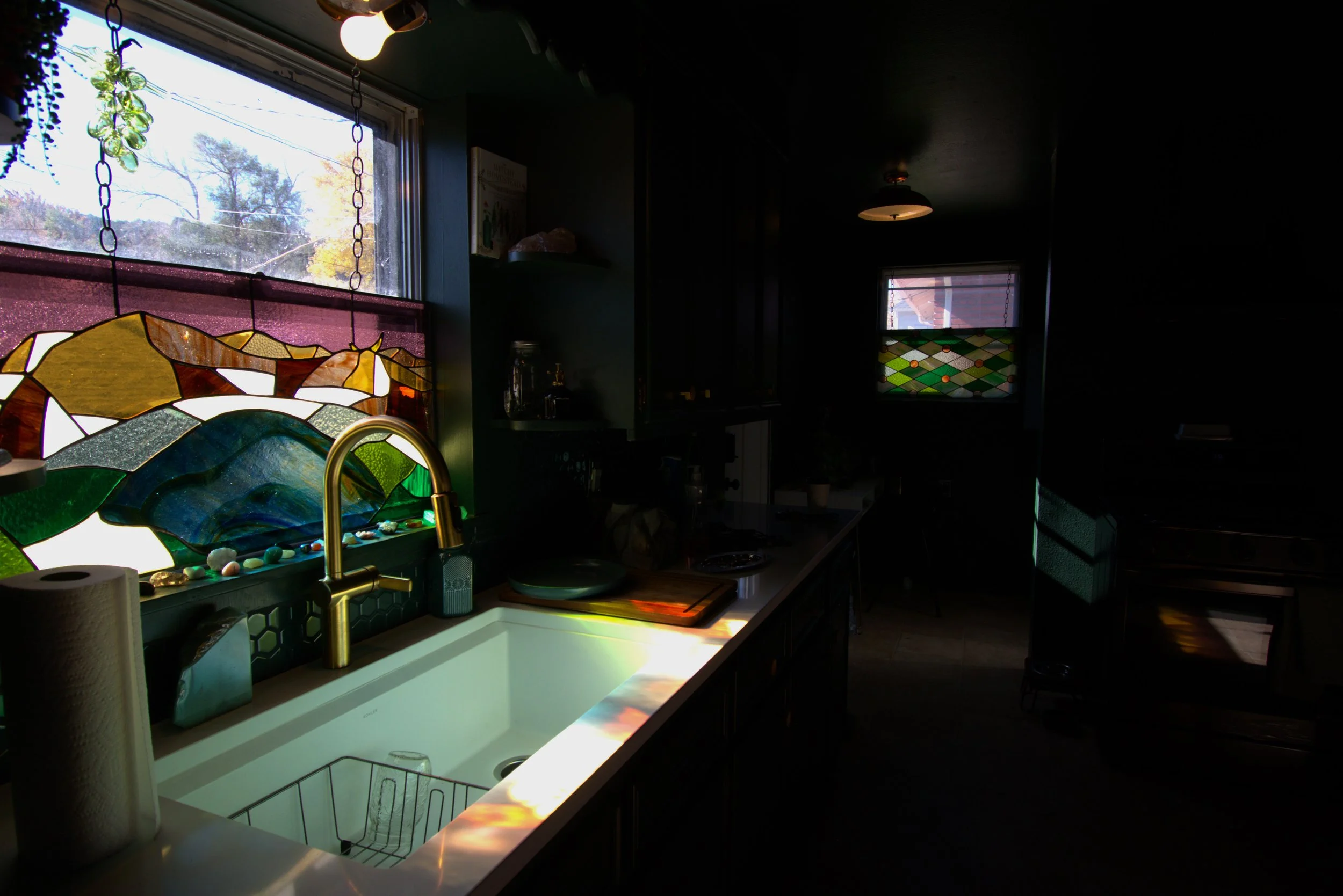 A kitchen with sunlight coming through stained glass windows, illuminating the sink area and creating colorful reflections on the countertop.