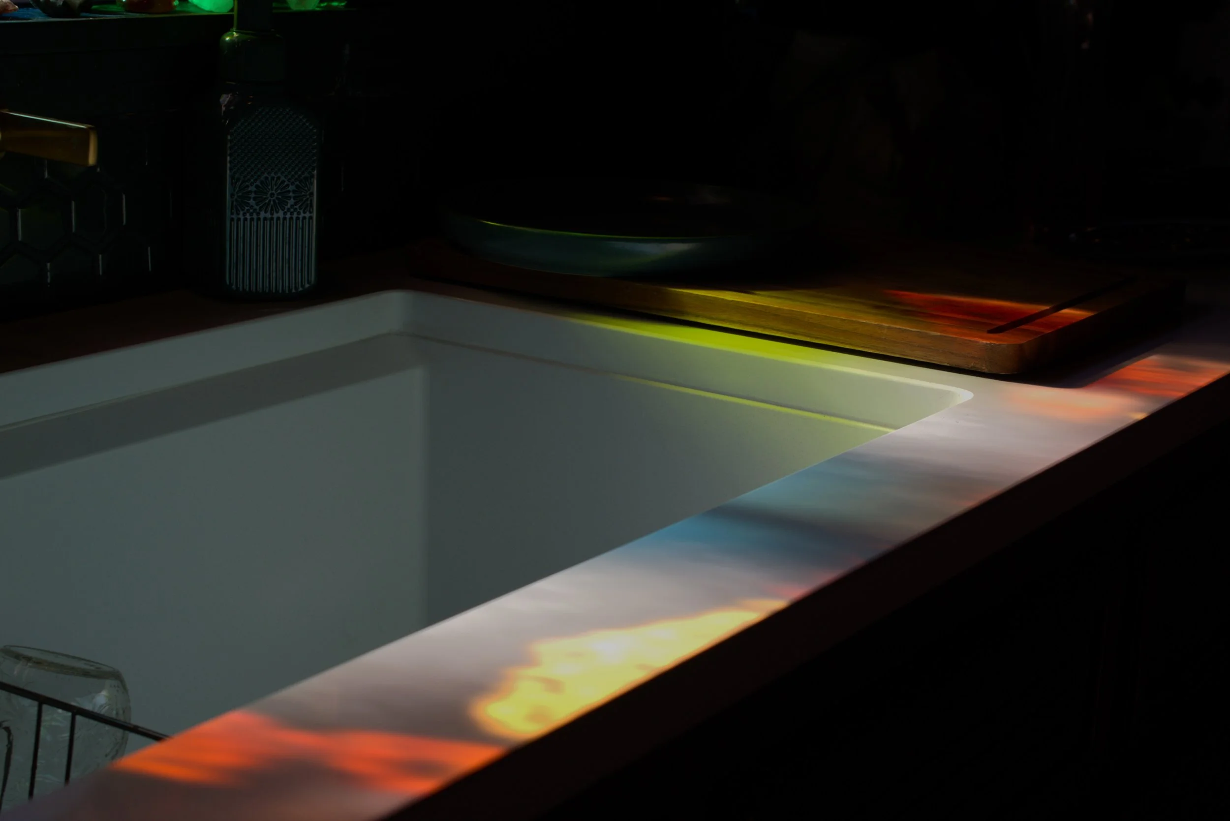 Empty white kitchen sink with colorful reflections on the countertop, a wooden cutting board, and some kitchen items in the background.