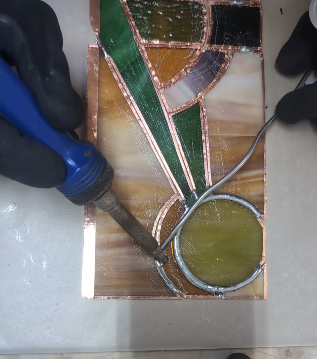 A stained glass art piece being soldered with a hot soldering iron, with sections of green, yellow, and black glass bordered by copper foil.
