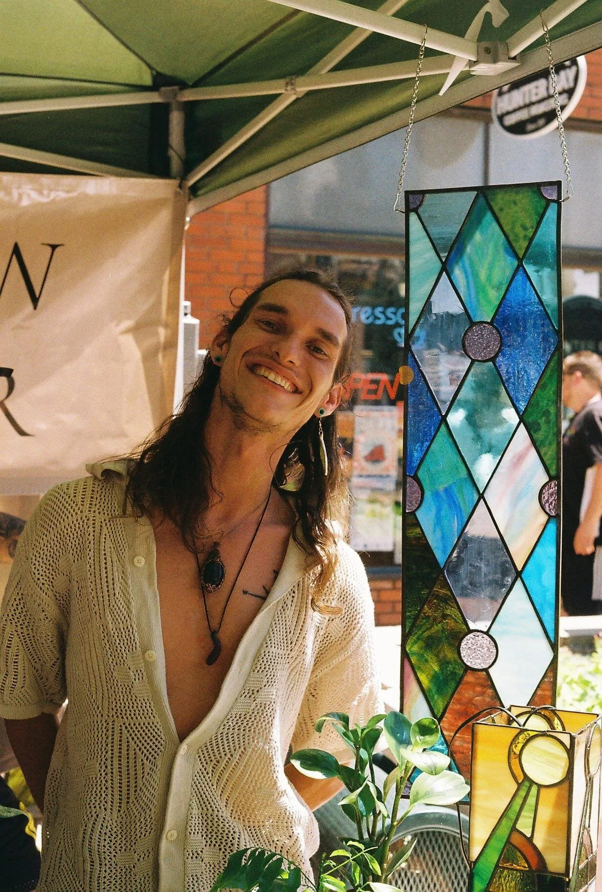 A smiling man with long brown hair, wearing a beige crochet shirt, necklace, and earrings, standing next to stained glass decorations at an outdoor market stall.