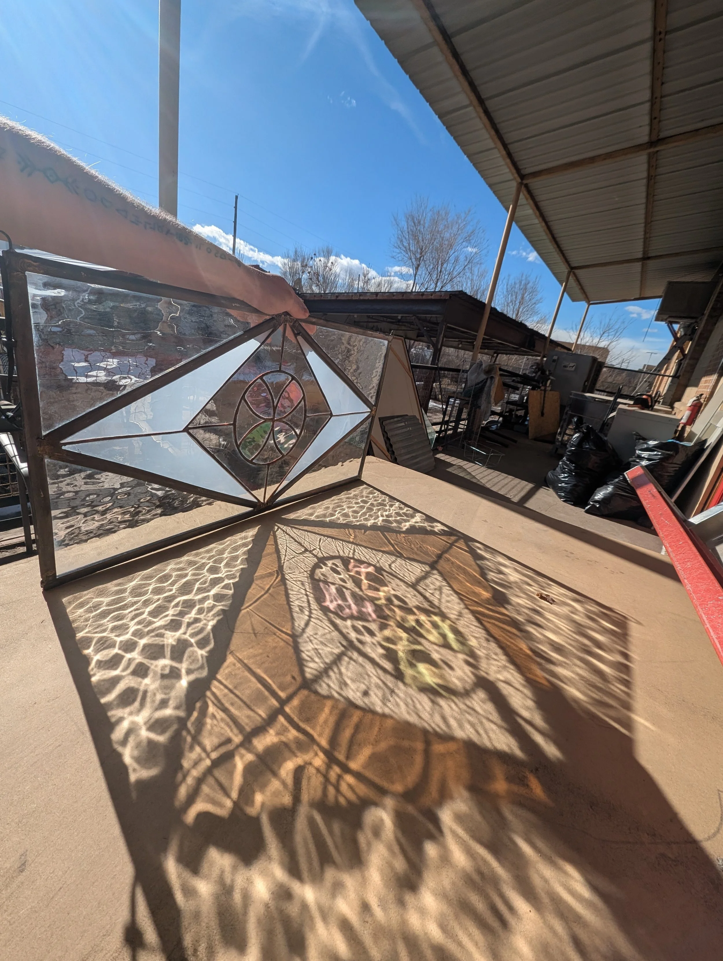 Sunlight passing through a stained glass window, casting colorful and intricate shadow patterns on a beige surface outdoors, under a metal roof.