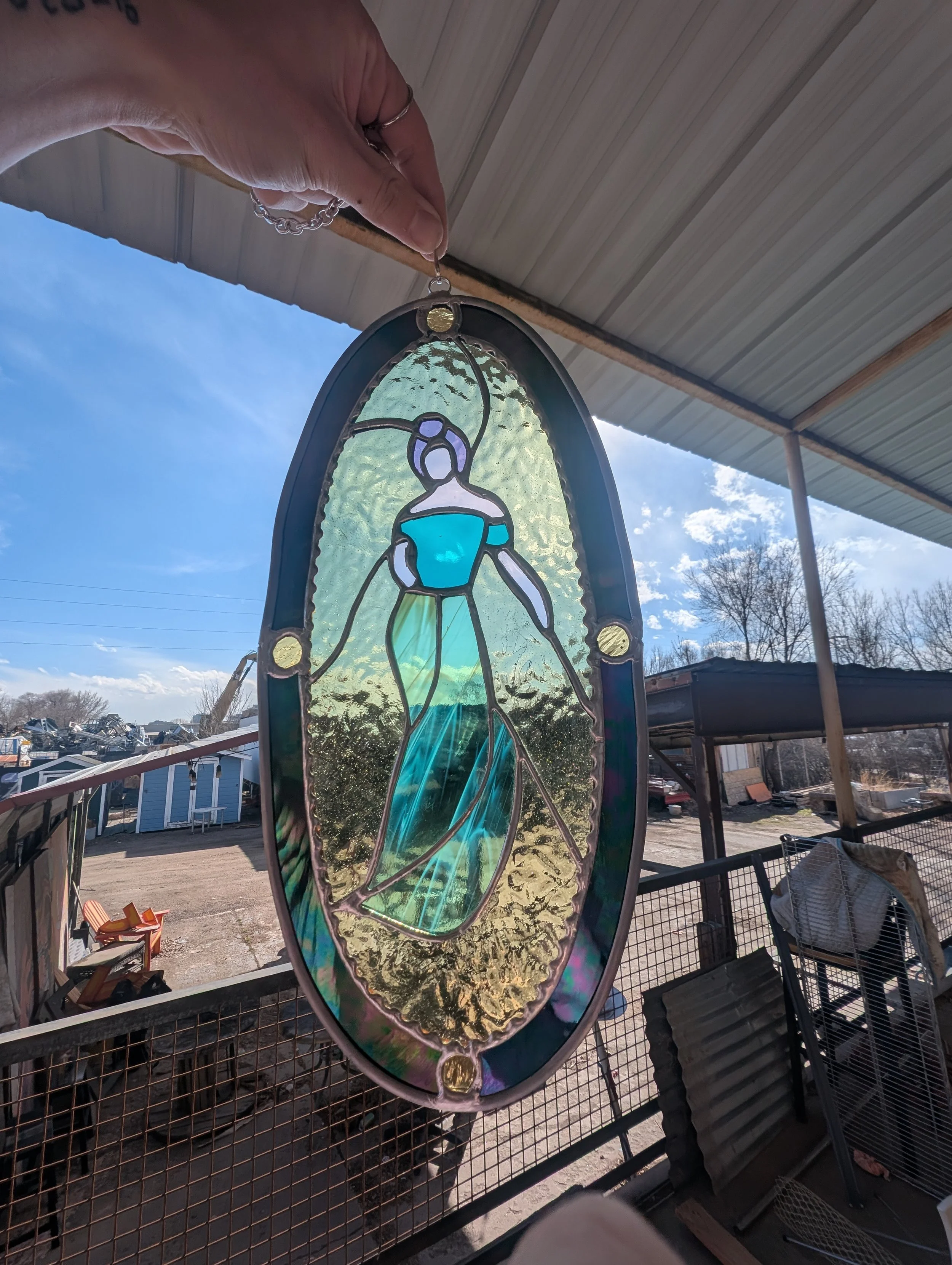 Colorful stained glass hanging ornament depicting a woman in a flowing dress, held up against a bright blue sky with trees and structures in the background.
