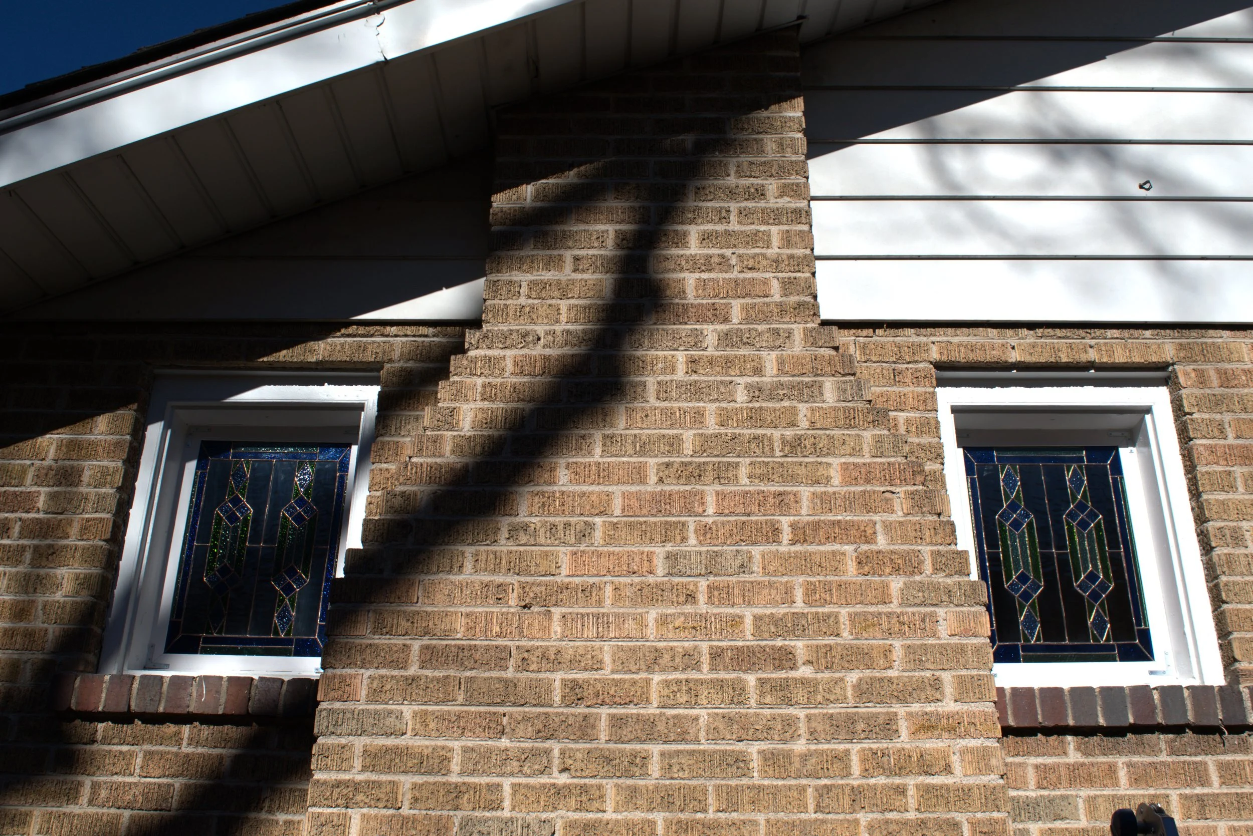 Close-up of brick house wall with two stained-glass windows, shadow of a tree branch, and part of the roofline.