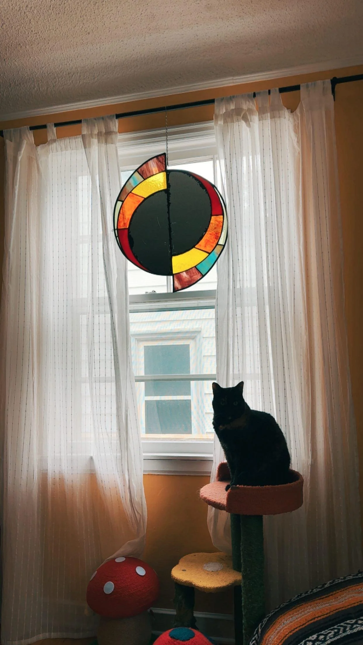 A black cat sitting on a pink perch of a multi-level cat tree in front of a window with white curtains. Above the cat tree is a colorful stained glass hanging lamp, and below the tree is a red and white mushroom-shaped decoration.