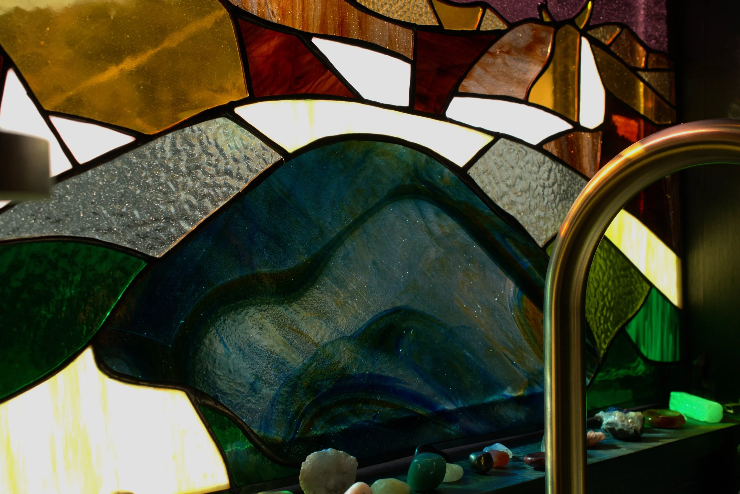Colorful stained glass window with various textured glass pieces and stones on a ledge underneath, featuring a prominent large blue-green piece and a gold-colored faucet to the right.