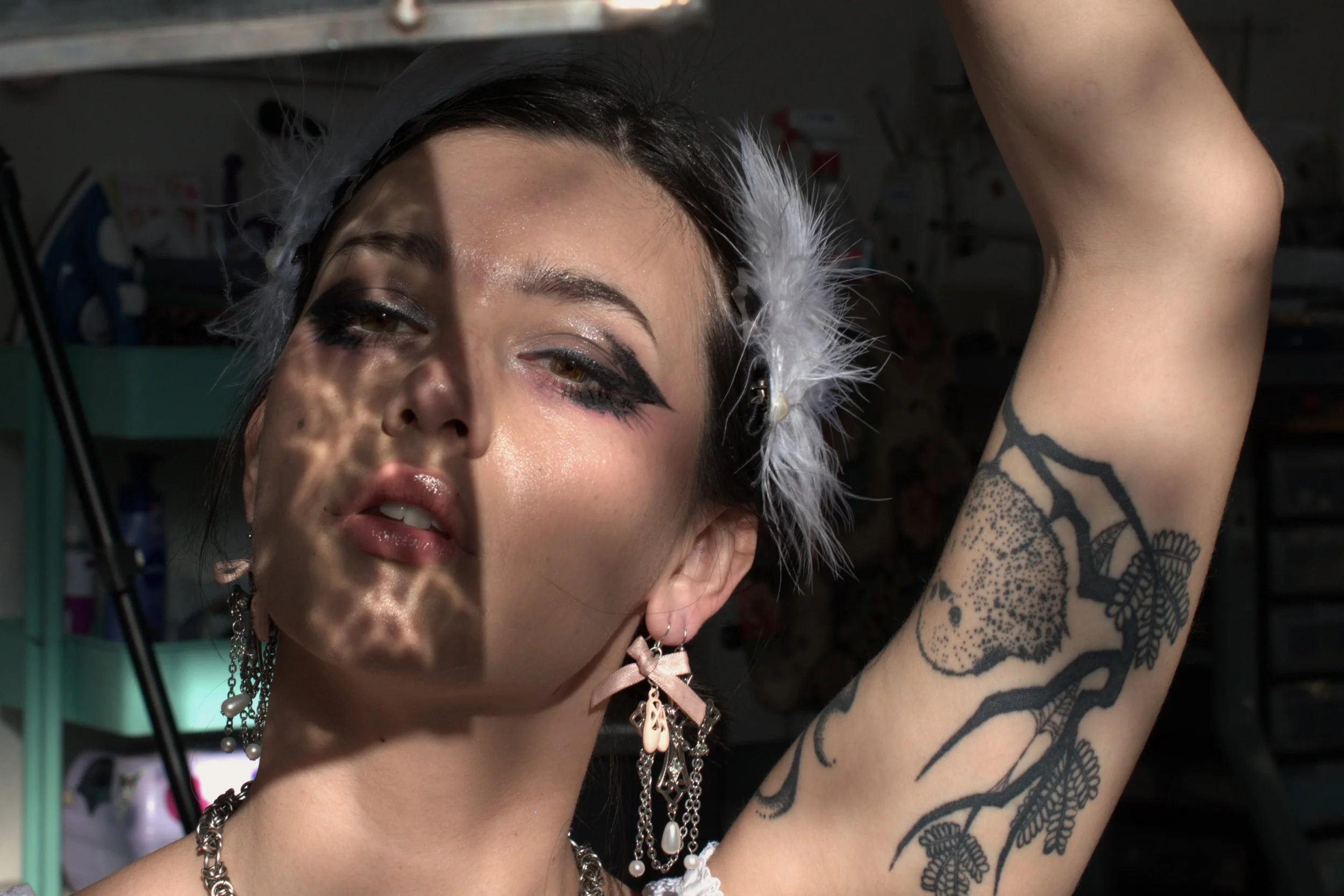 A woman with dark makeup and a feathered headpiece, partially shadowed and illuminated by light that creates a patterned shadow on her face, showing her with a tattoo on her arm, earrings, and a necklace.