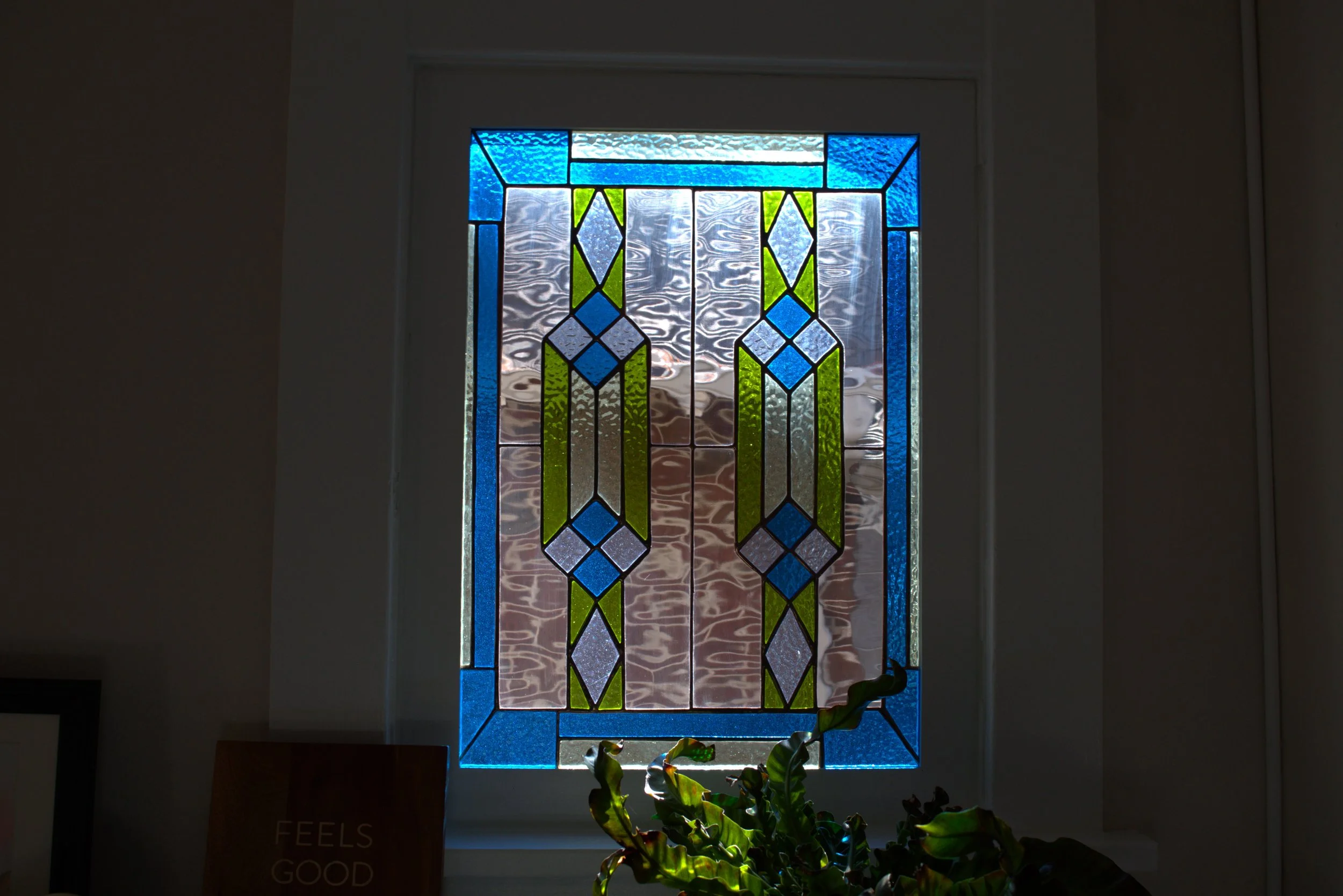 Colorful stained glass window with geometric patterns in blue, green, and clear glass, with a potted plant and a small sign that reads 'FEELS GOOD' beneath it.
