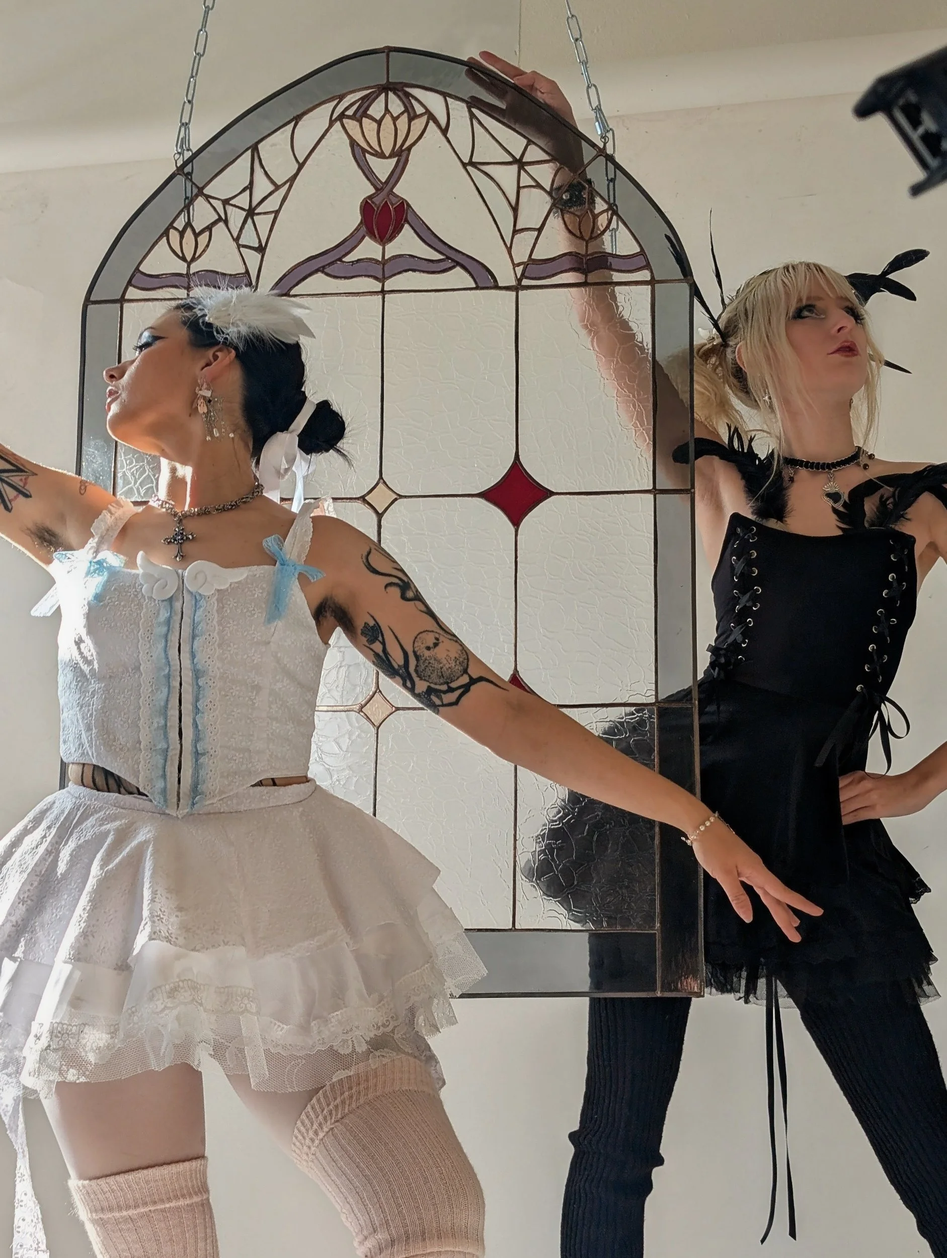 Two women dressed in fantasy or cosplay costumes stand in front of a stained glass mirror. One wears a white dress with lace, feathers, and accessories, with tattoos on her arm. The other wears a black lace-up outfit with feathered headbands and blon