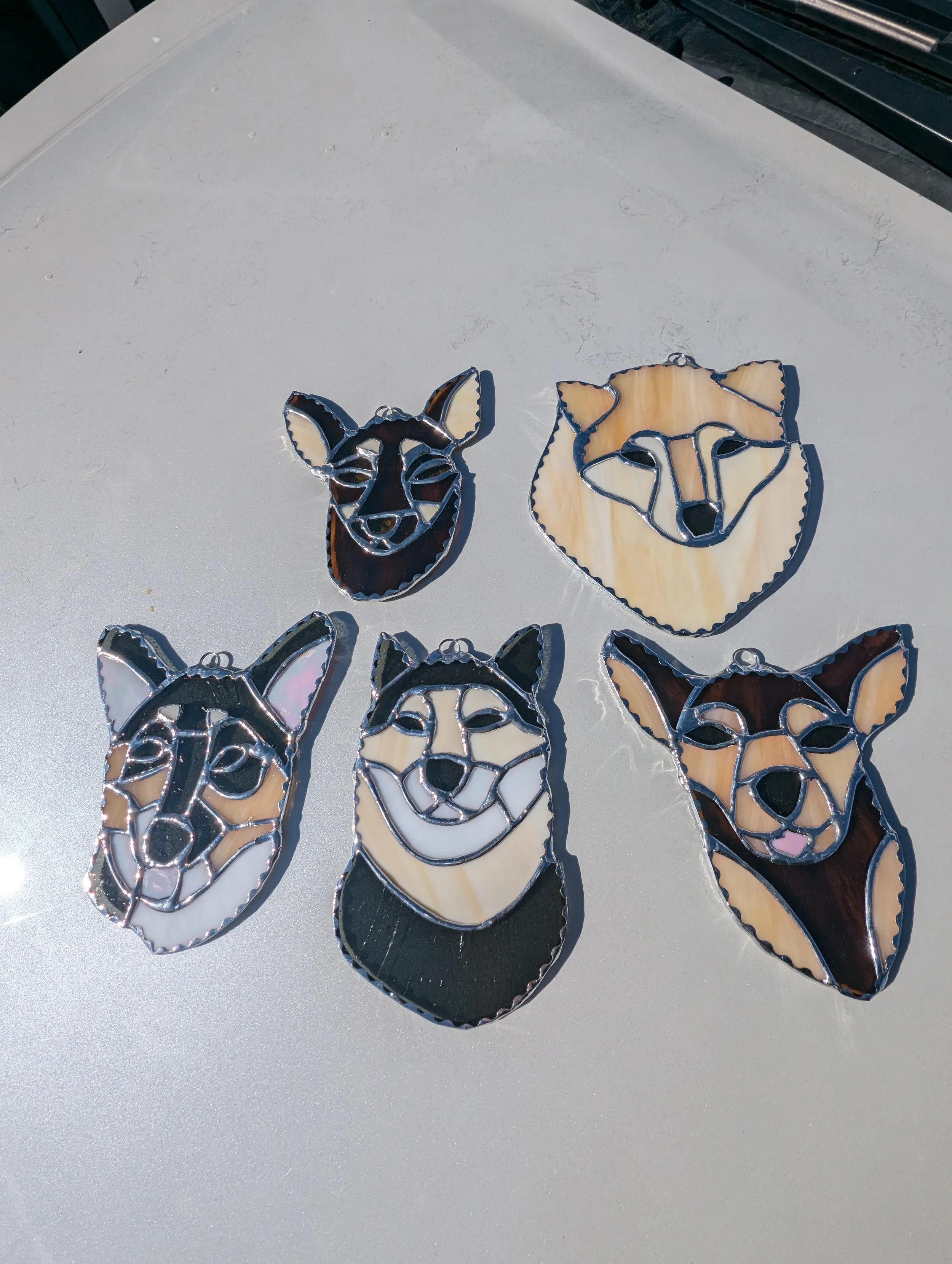Five stained glass dog faces, each with different designs and colors, arranged on a light surface outdoors.