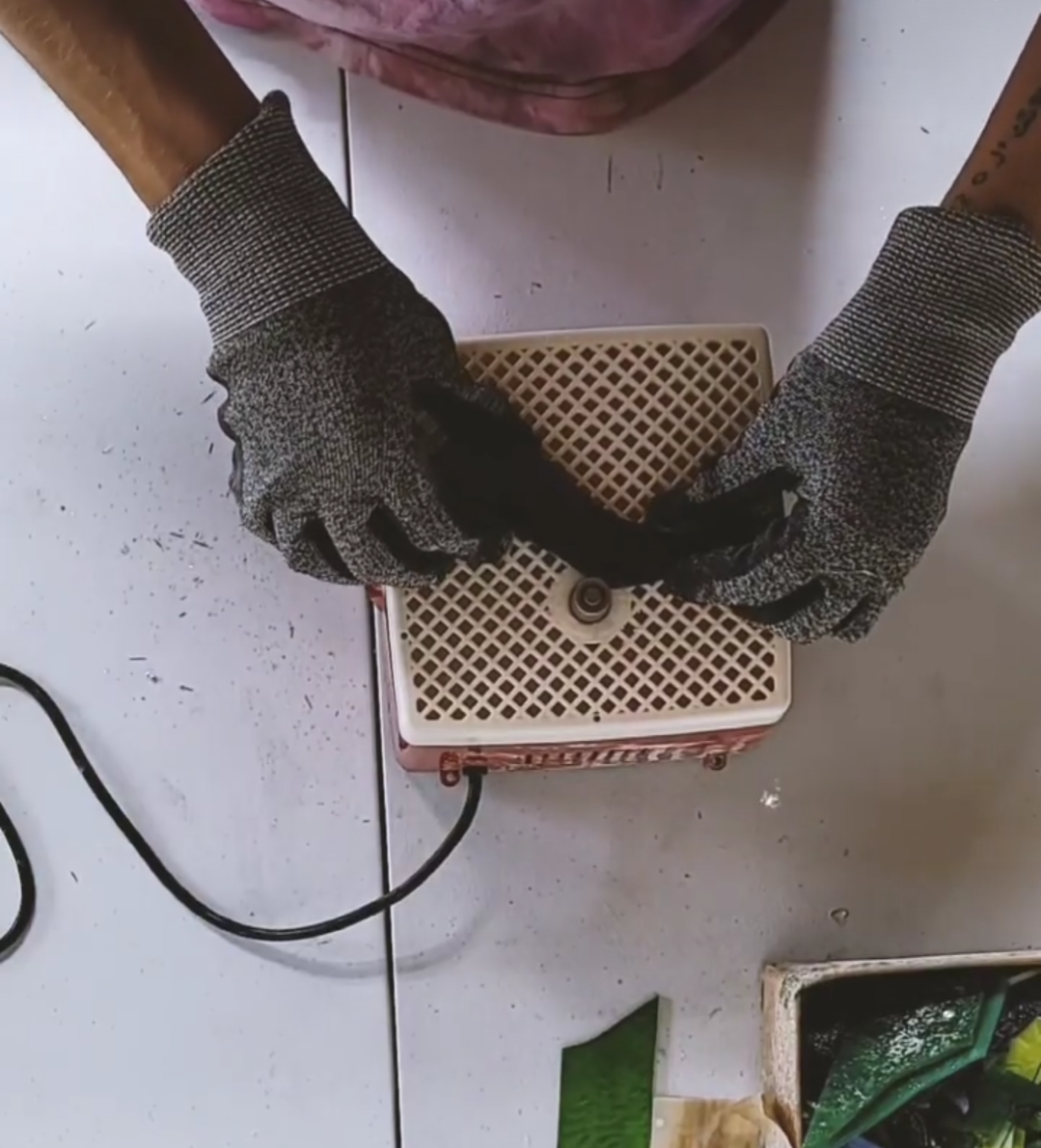 Person wearing black and gray gloves handling a small electrical component or device over a white surface, with a small orange and white device and a black cord nearby.