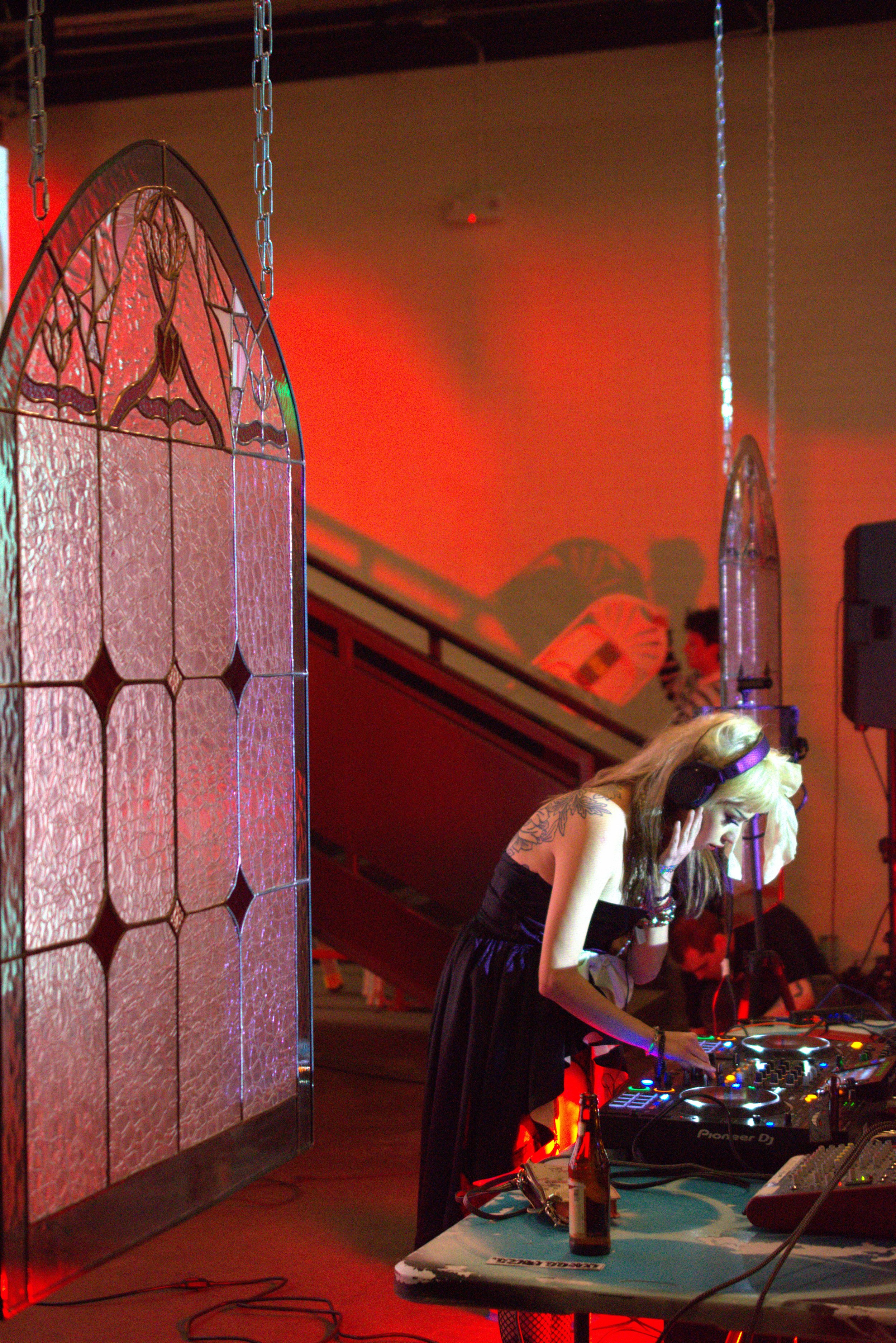 A female DJ with long blonde hair, wearing headphones and a black dress, is DJing at an event with red lighting, a stained glass window, and a beer bottle on a table nearby.
