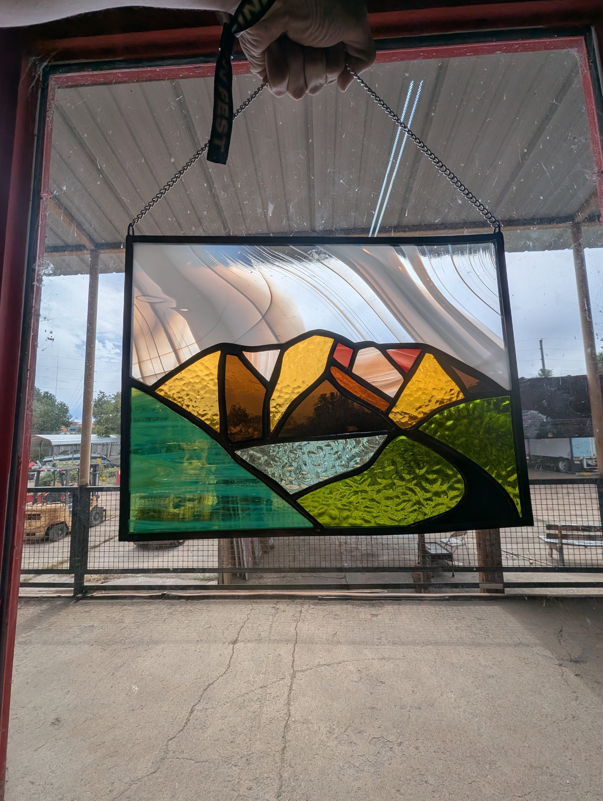 Colorful stained glass window depicting a mountain landscape with green fields, rocky mountains, and a cloudy sky.