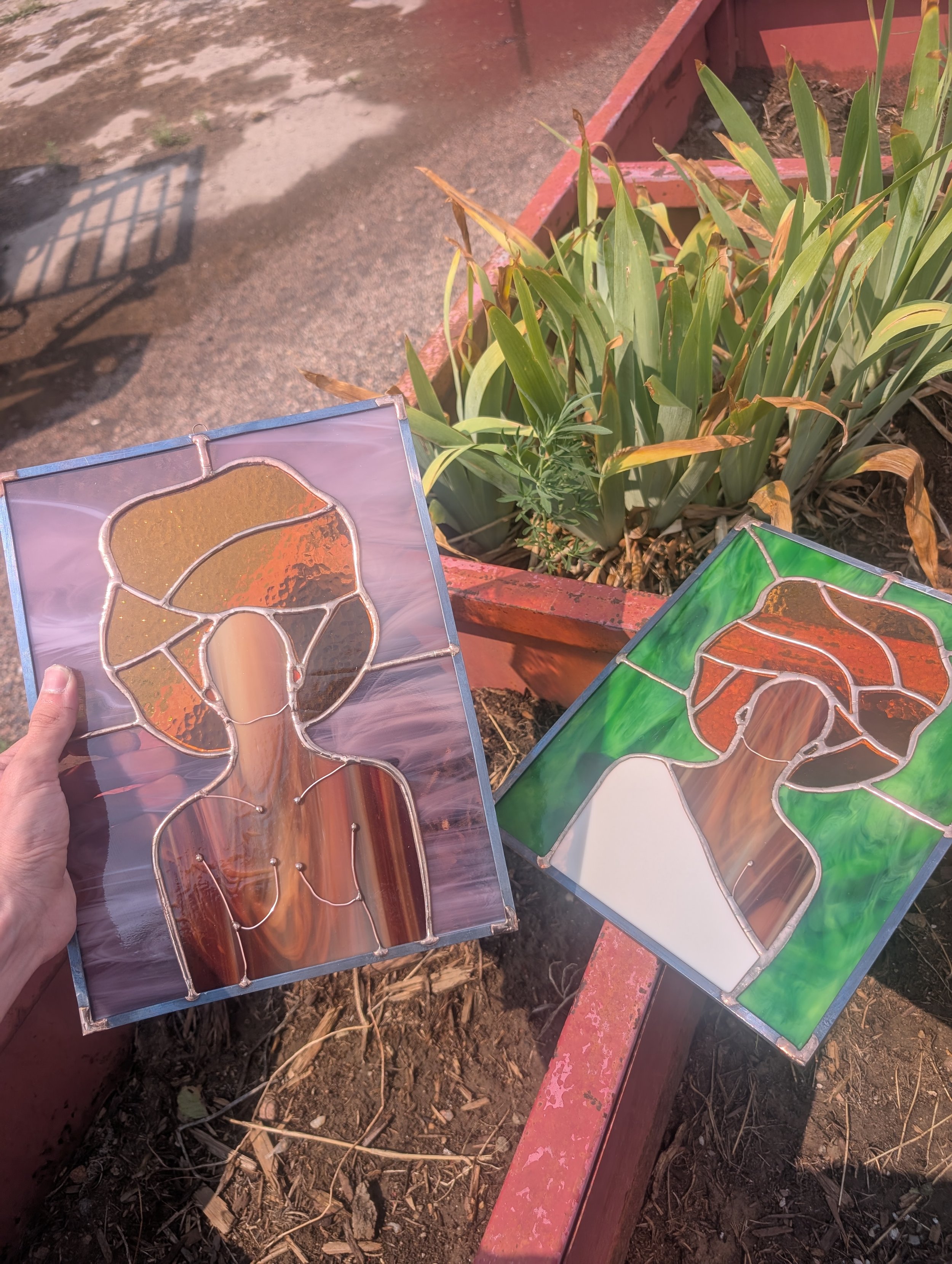 Two pieces of stained glass artwork depicting human faces with broccoli-like hair, one with a background of swirling purples and browns, the other with a green background, placed on a red planter box outdoors next to a plant.