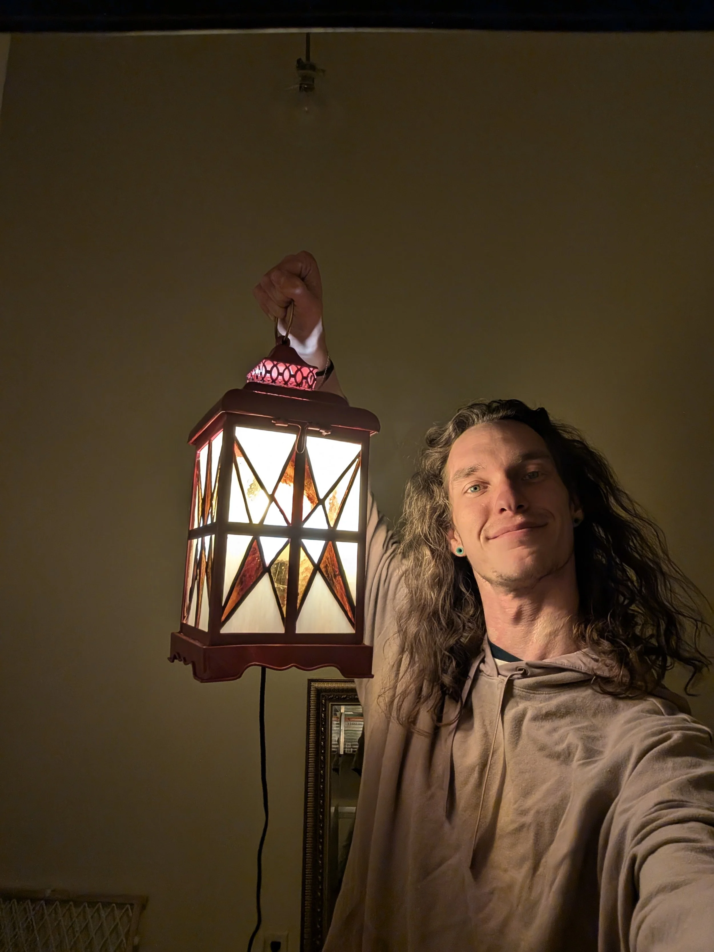 A person with long, curly hair and a light-colored hoodie holding a hanging lantern, smiling at the camera in an indoor setting.