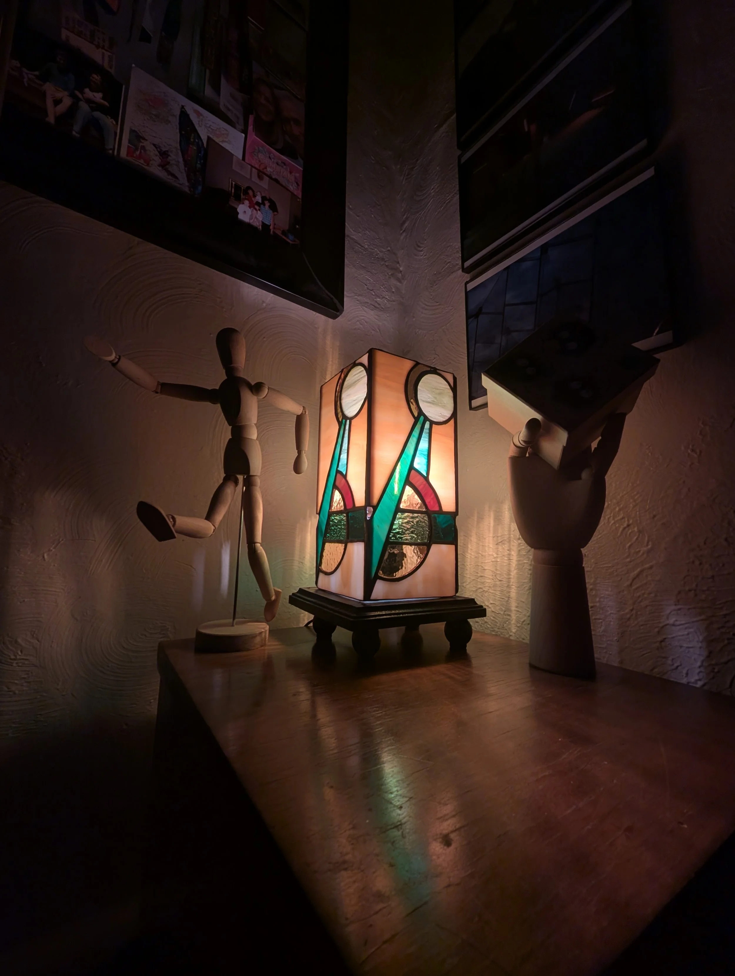A wooden artist mannequin posed as if running, a colorful stained glass table lamp, and a white ceramic hand sculpture holding a black box holding electronic devices, all on a wooden surface against a textured wall with mounted TV screens and photos.