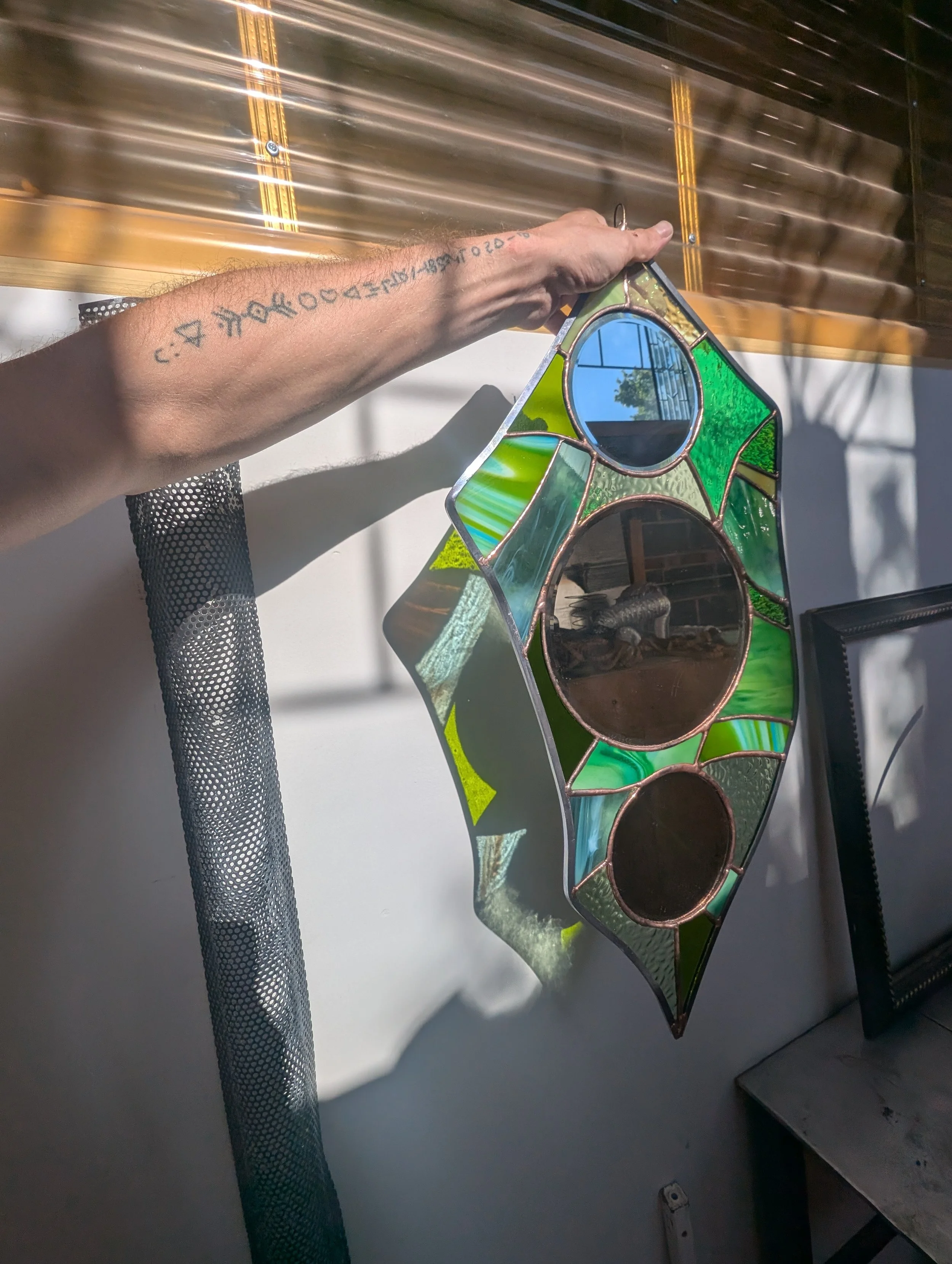 Person hanging stained glass window on a white wall, with sunglasses reflected in the glass. The person's arm has tattoos.
