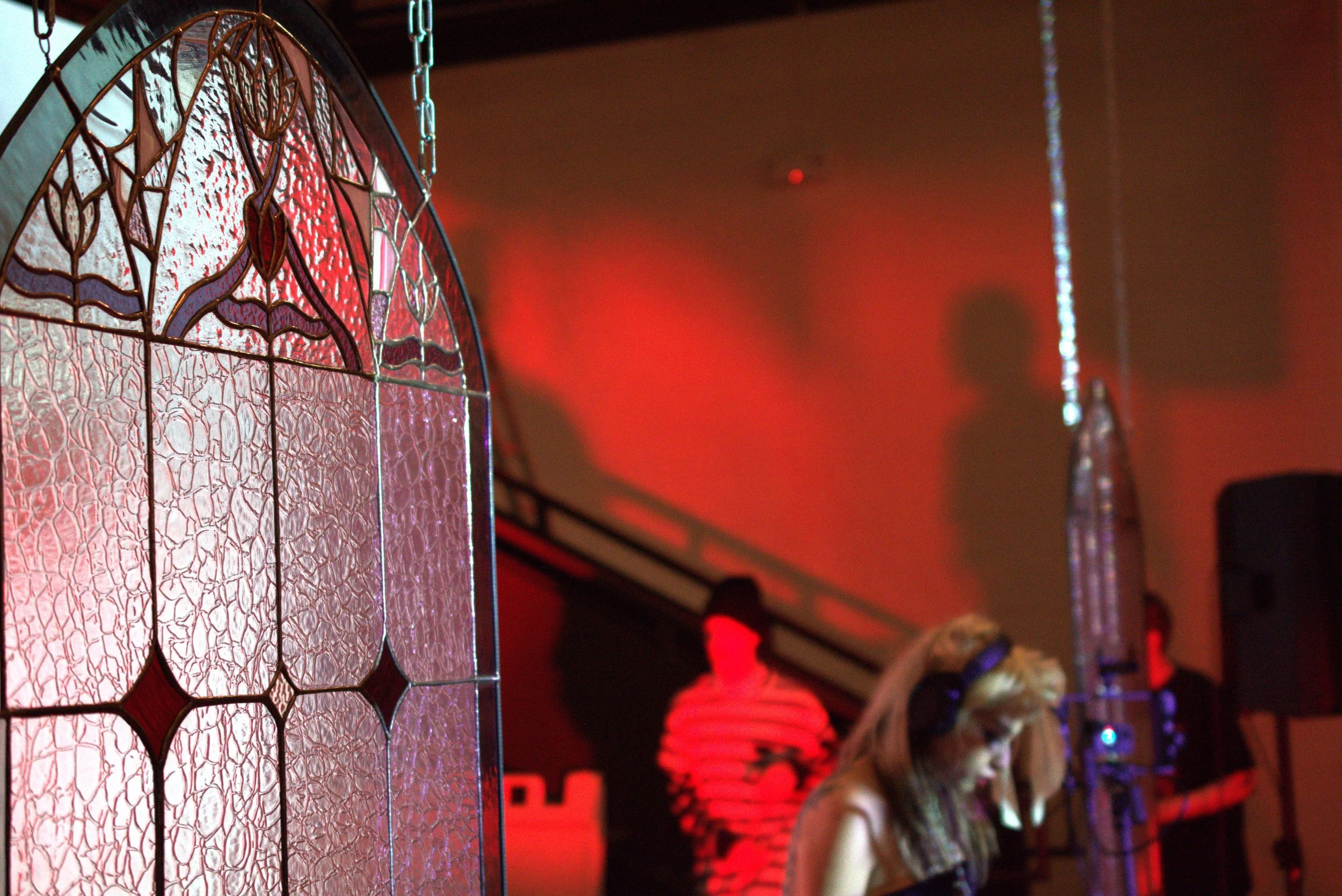 A stained glass window with a decorative pattern in the foreground, and a woman wearing headphones looking down in the background at a party or event with red lighting.
