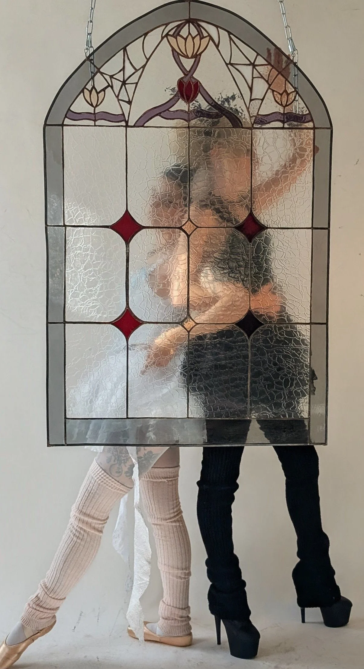 Two dancers, a ballerina in a tutu and pointe shoes on the left, and a woman in high heels and black pants, are reflected in a stained glass mirror with a floral design. They are posing with arms raised and one leg extended.