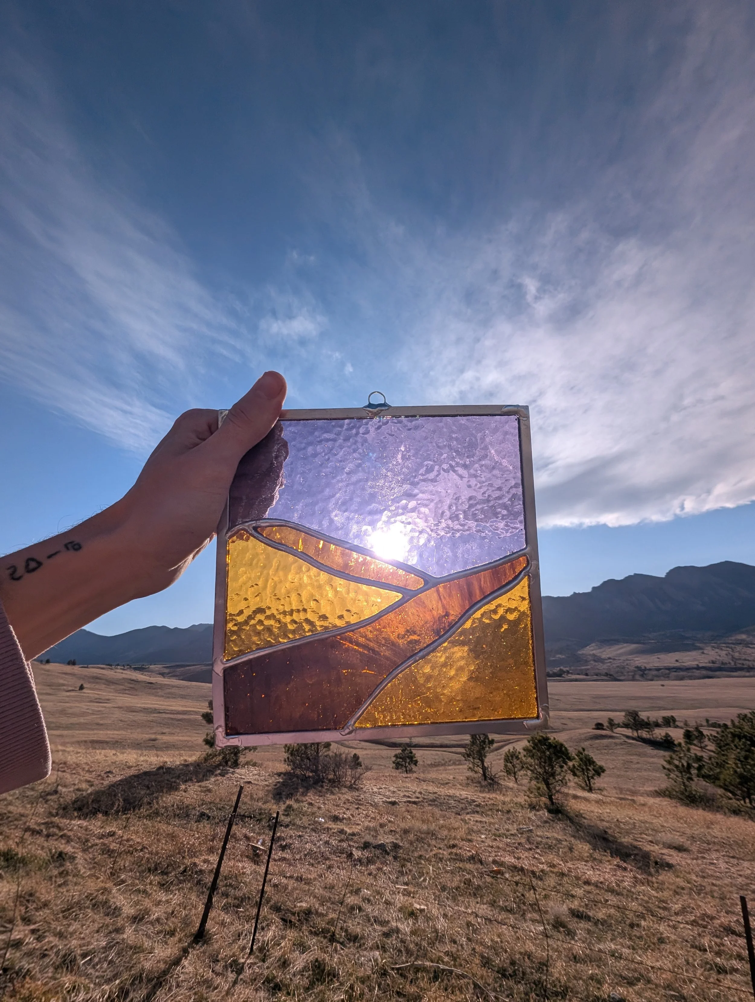 A person holding a stained glass panel with a landscape design against a background of rolling hills, trees, mountains, and a blue sky with wispy clouds.