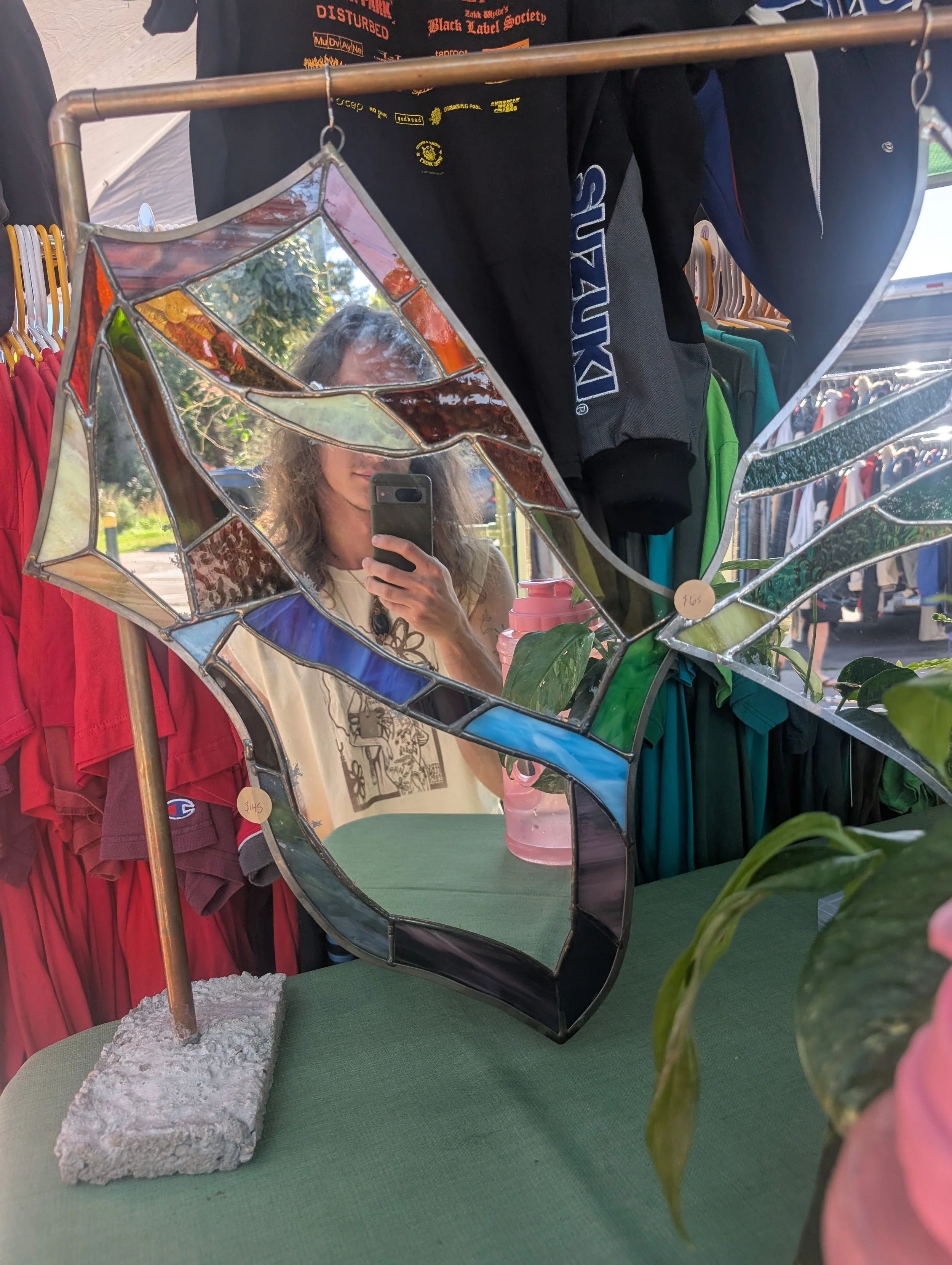 A decorative stained glass mirror reflecting a woman taking a selfie with her phone. The mirror is shaped like a butterfly with colorful stained glass sections. The scene is set at an outdoor market with clothing hanging in the background.