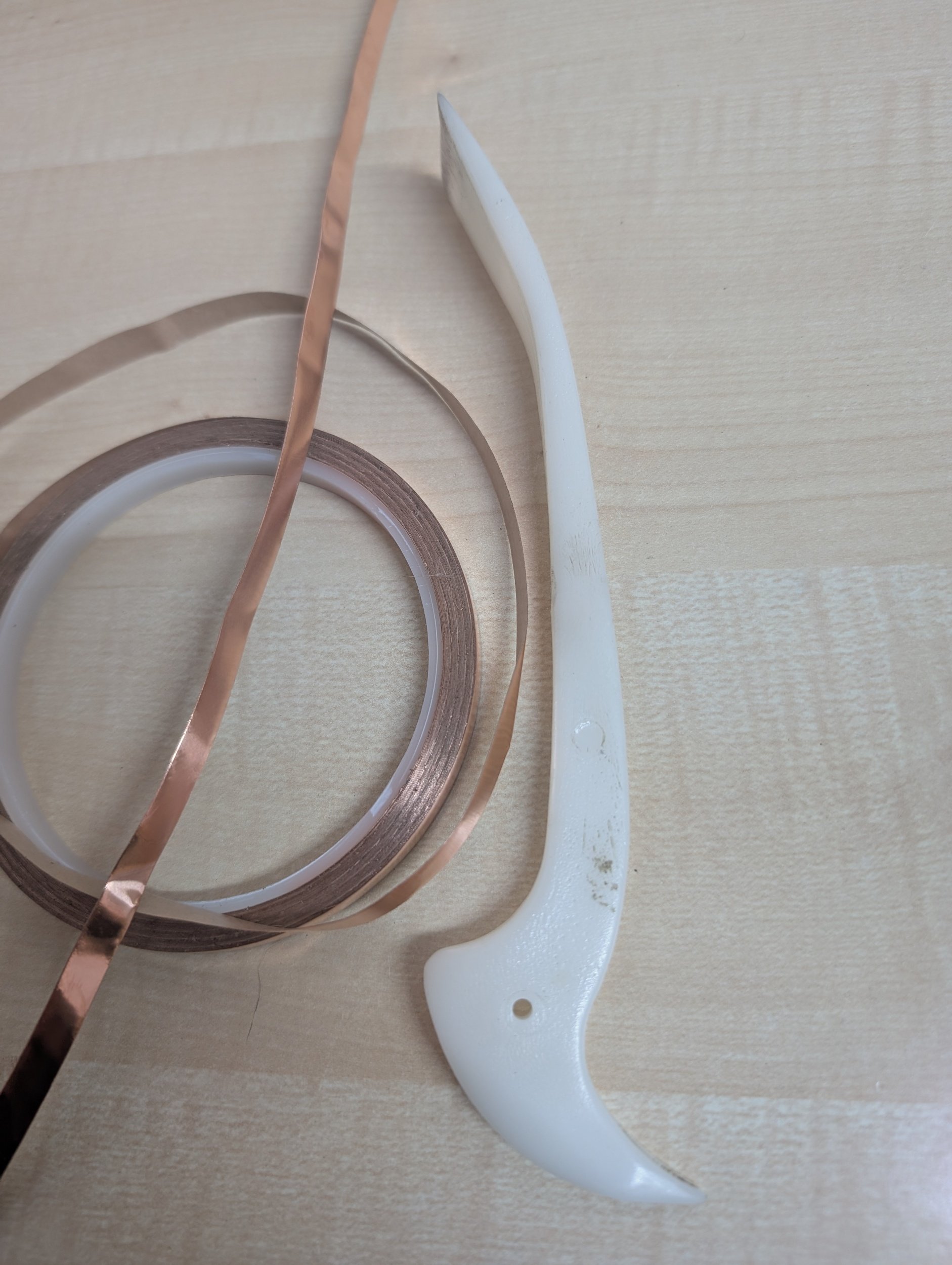 A white plastic shark-shaped tool, a copper-colored strip, and some circular tapes on a wooden surface.