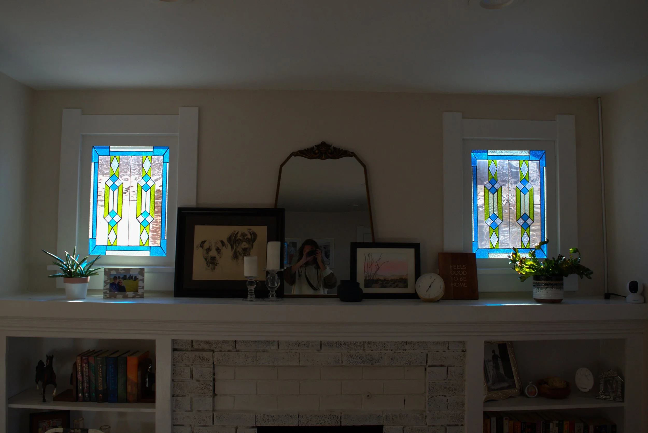 Interior of a living room with a white brick fireplace, two stained glass windows, a mirror, framed artwork, potted plants, decorative items, and shelves with books.