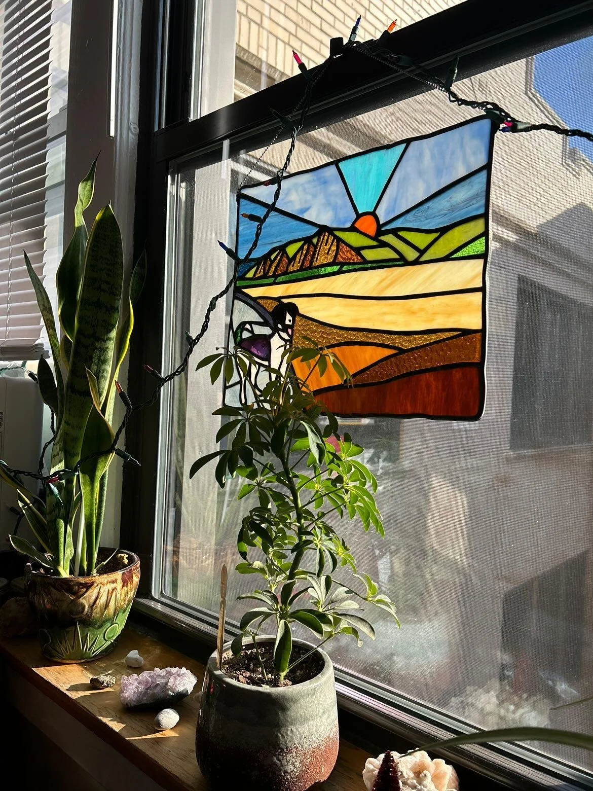 Sunset or sunrise stained glass window hanging, with a landscape scene including mountains, fields, and a bird, hanging in front of a window with house and sky visible outside. Potted plants and decorative stones on a windowsill.