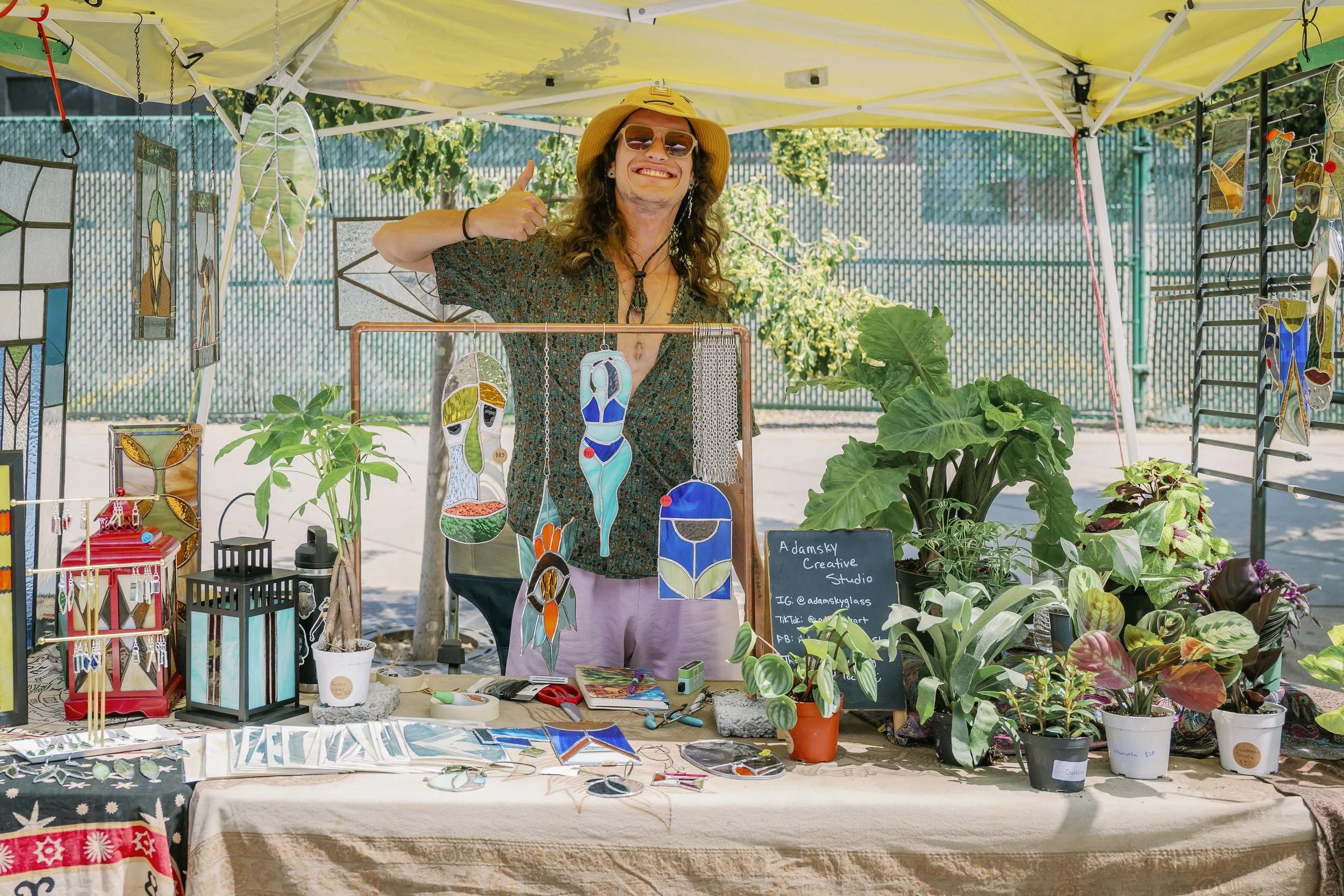 A man with long hair, wearing a beige hat, sunglasses, and patterned shirt, standing at an outdoor art booth. He is smiling and giving a thumbs-up. The booth displays colorful stained glass art, jewelry, and potted plants, with a sign indicating it i