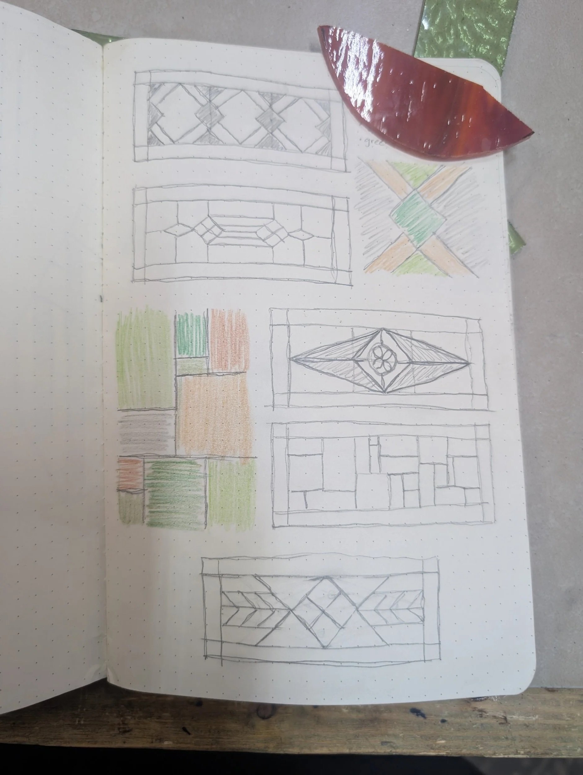 Sketchbook page with various stained glass window designs, some colored with green, orange, and beige, alongside a red shell and green rectangular pieces, possibly for reference.