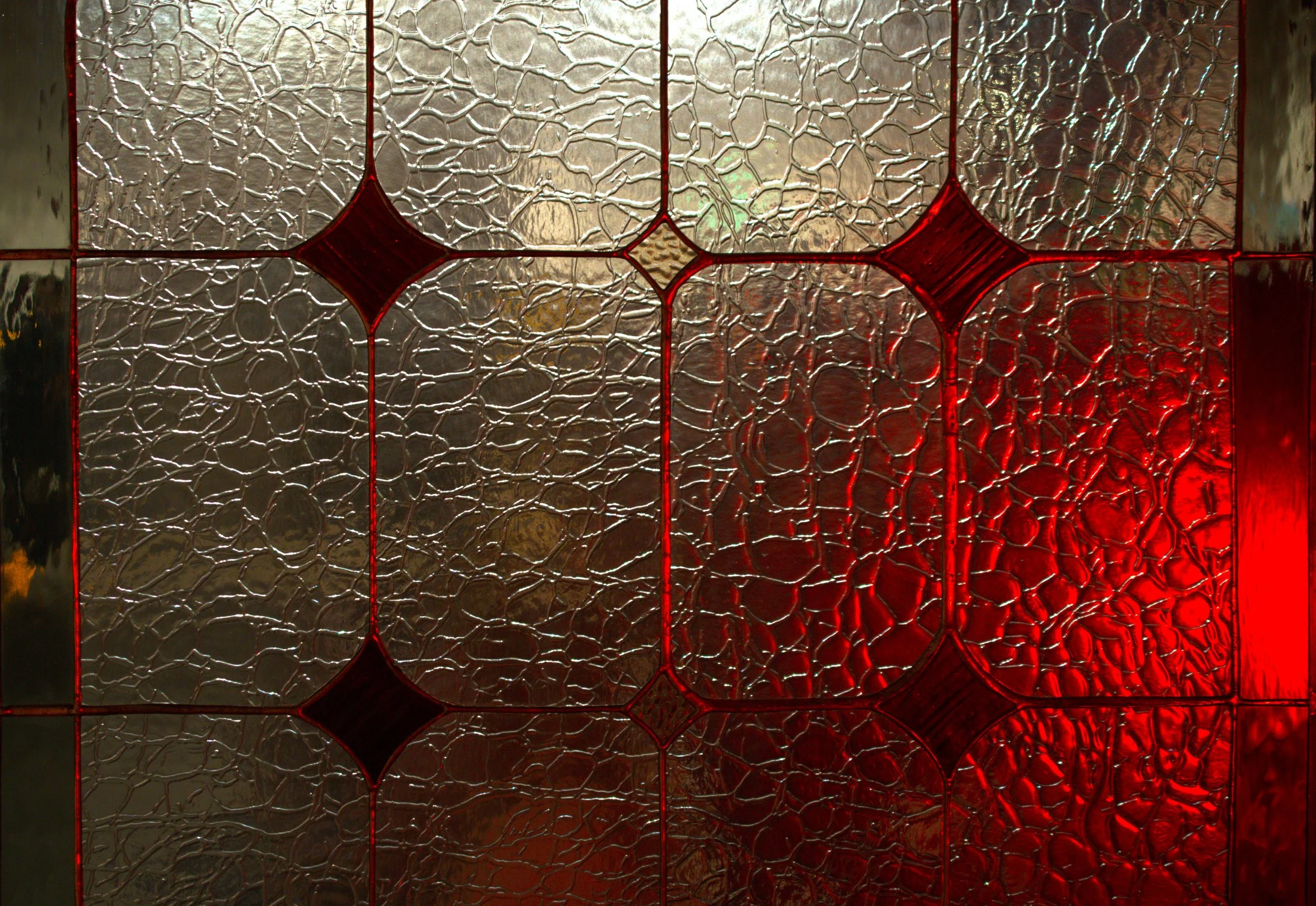 A close-up of textured glass stained with red and metallic elements, organized in a geometric pattern.