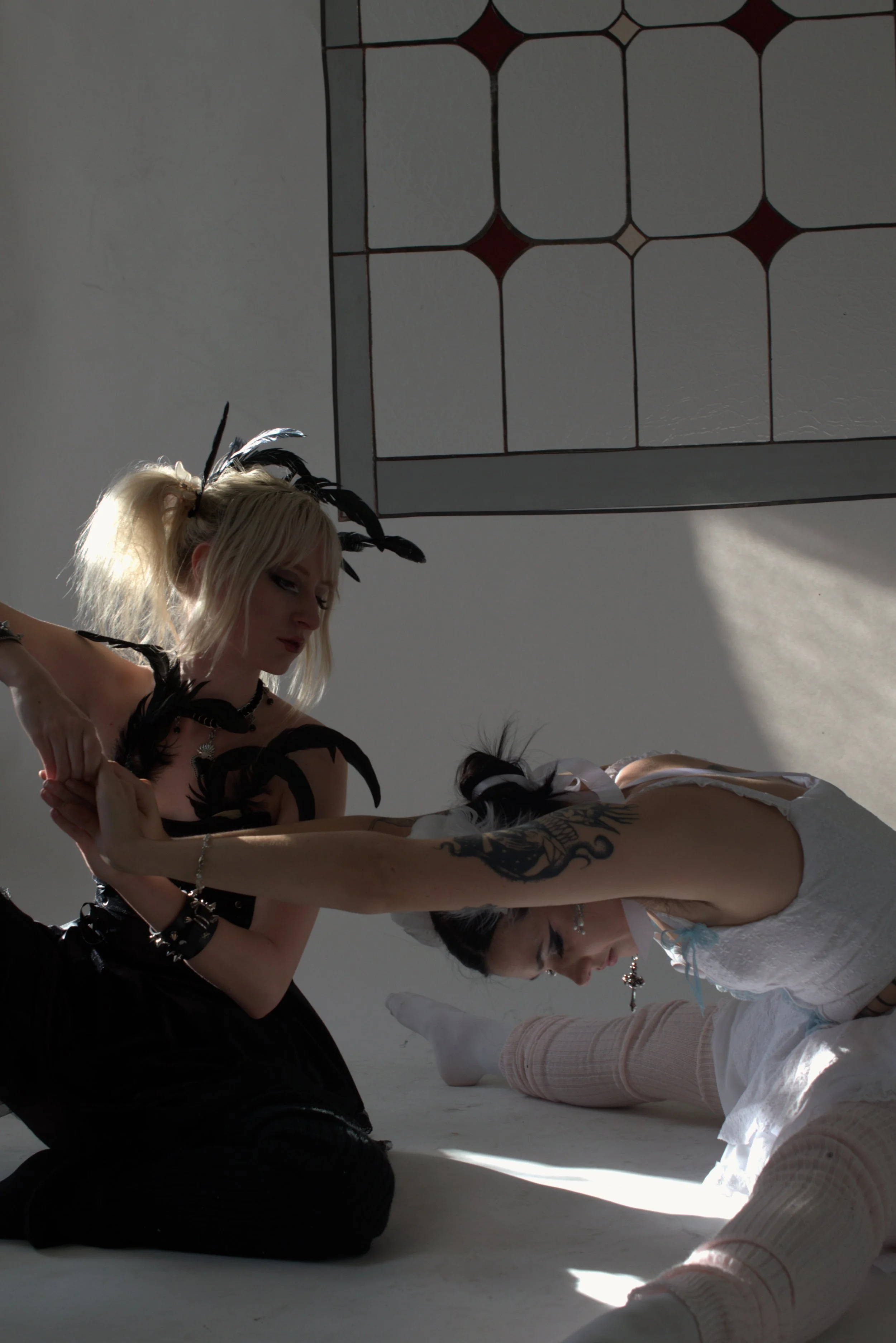 Two young women engaged in a playful wrestling or dance on the floor near a wall with a stained glass window, one with blonde hair and feathers in her hair, the other with dark hair and tattoos, in a well-lit room with sunlight streaming in.