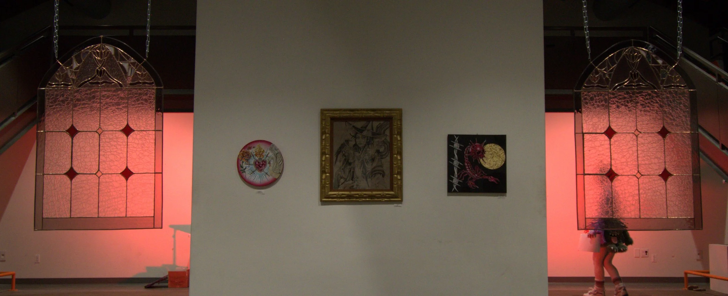 Art gallery featuring three pieces of artwork on a white wall, flanked by two large stained glass windows with red lighting, staircase on both sides, and a person with a floral hat sitting on a bench in the corner.