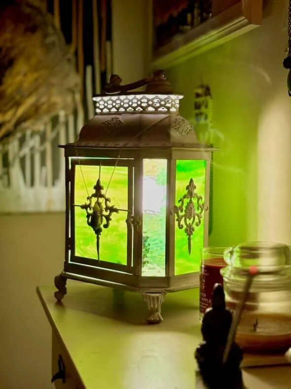 An ornate decorative lantern with glass panels that have green swirling designs, placed on a table in a room with a green wall and various objects nearby.