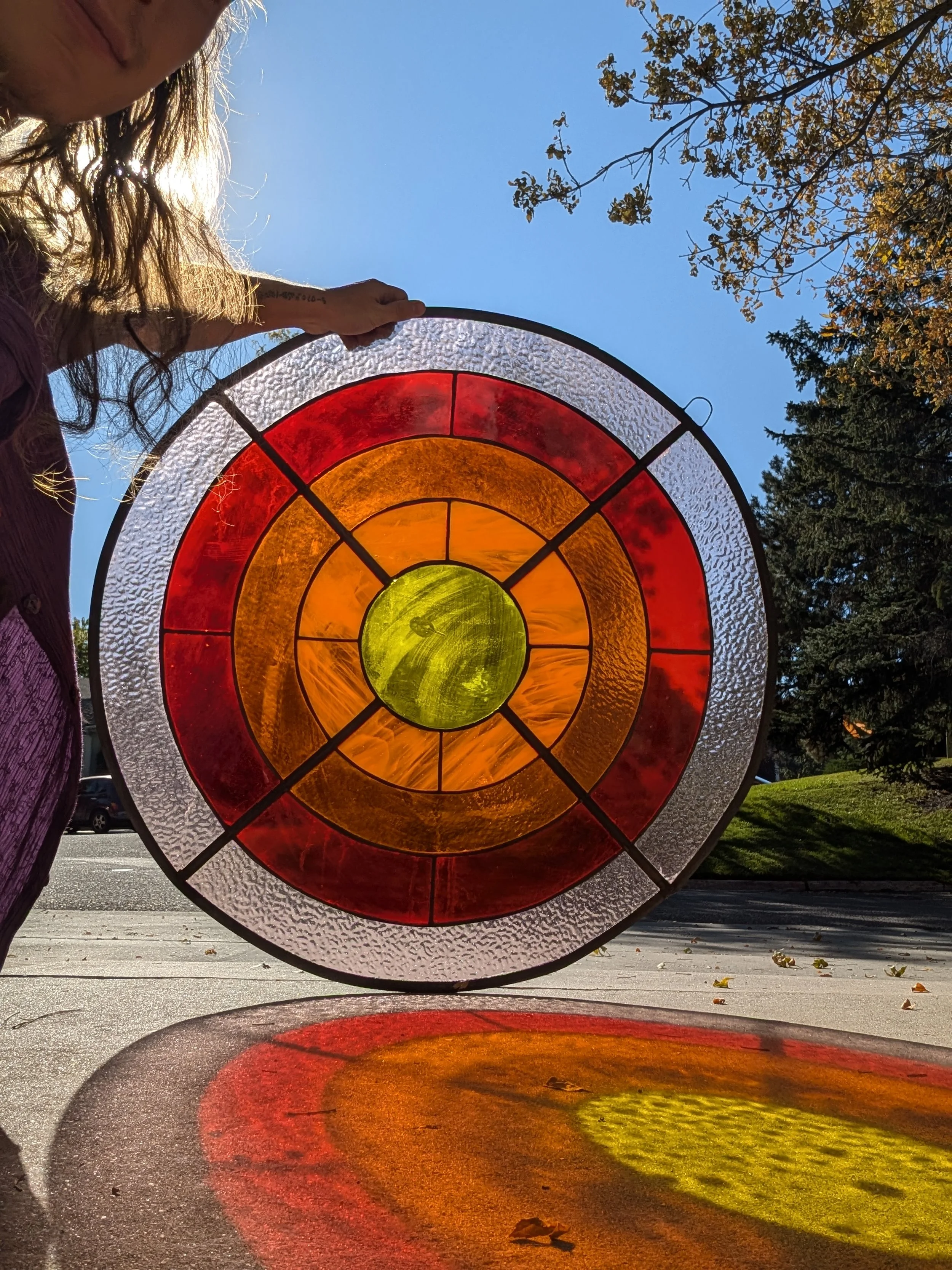 A person holding a stained glass circular artwork outdoors, with sunlight shining through it, casting colored shadows on the ground.
