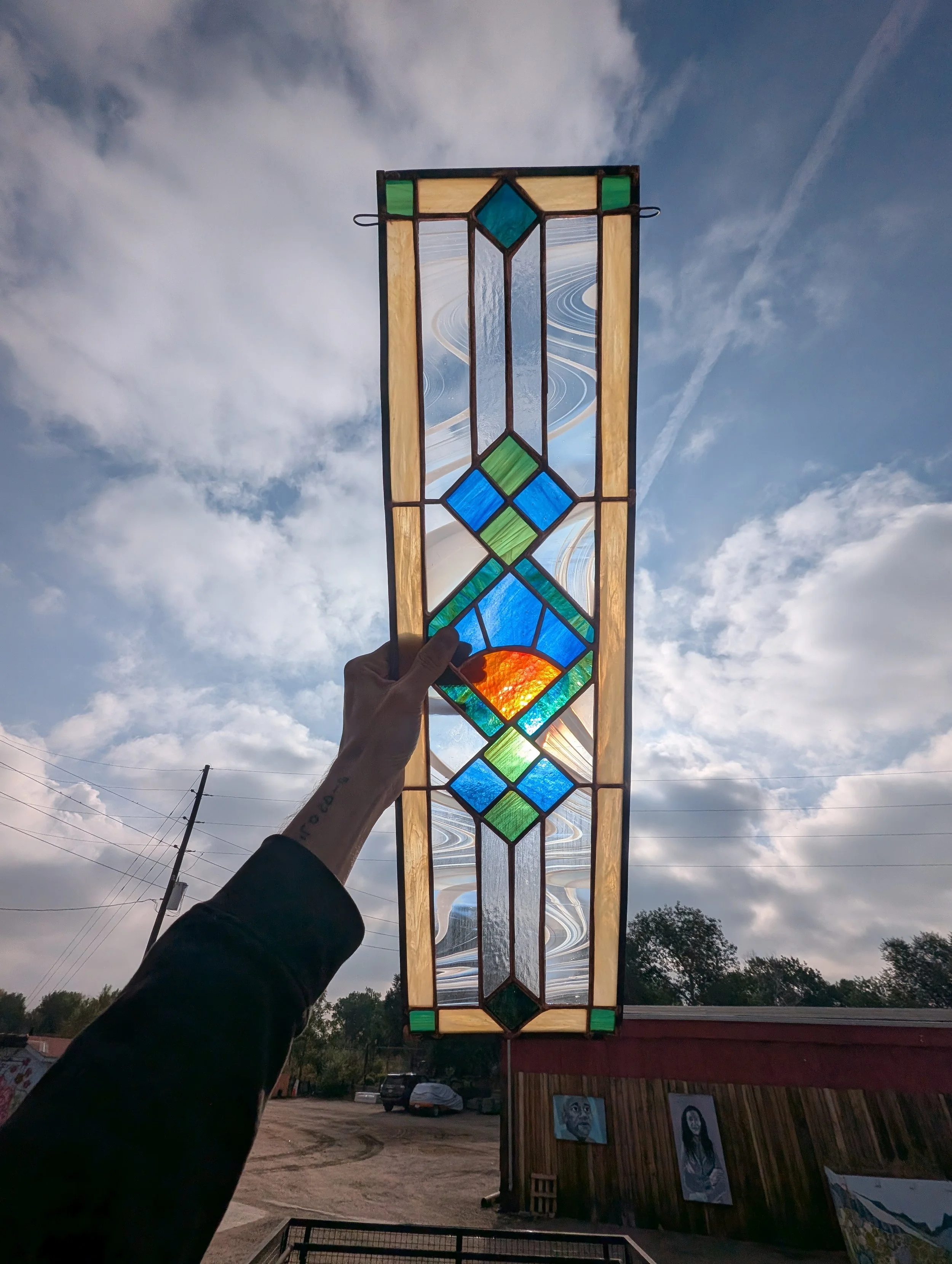 Person holding a stained glass window against the sky, with clouds, trees, and parked cars in the background.