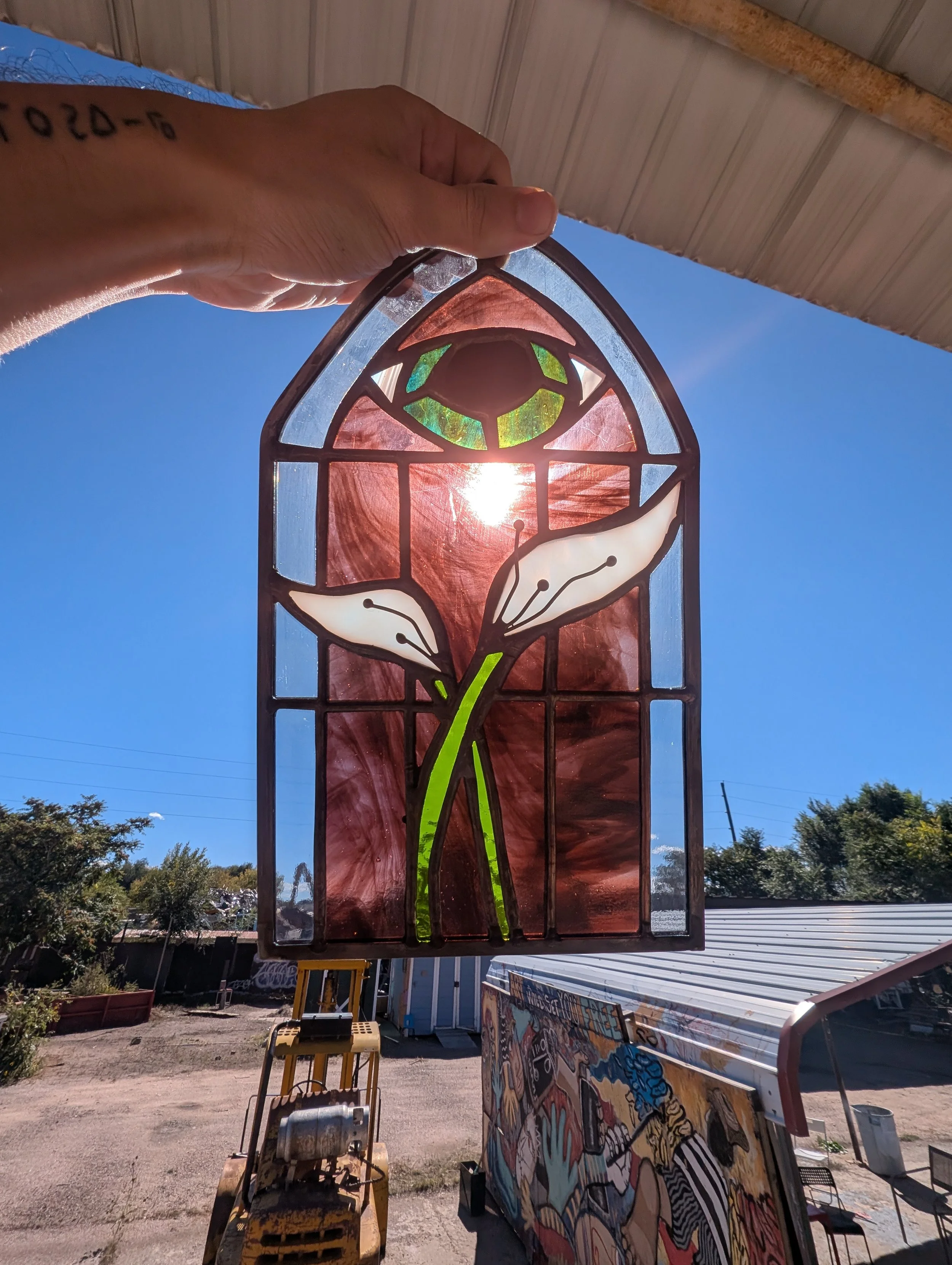 Hand holding a stained glass window with a flower design, sunlight shining through, outdoor setting with equipment and graffiti artwork in the background.