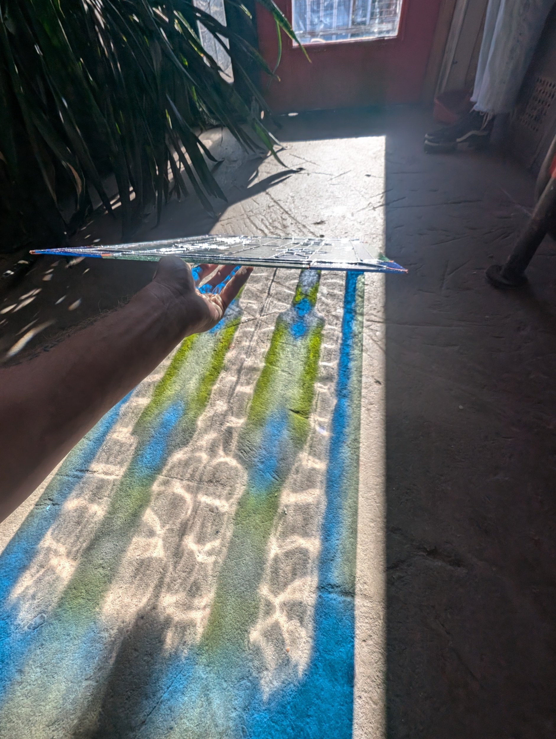 A hand holding a broken piece of glass reflects sunlight, casting colorful shadows on a textured concrete floor. In the background, there is a door letting in sunlight, plants, and some household items.