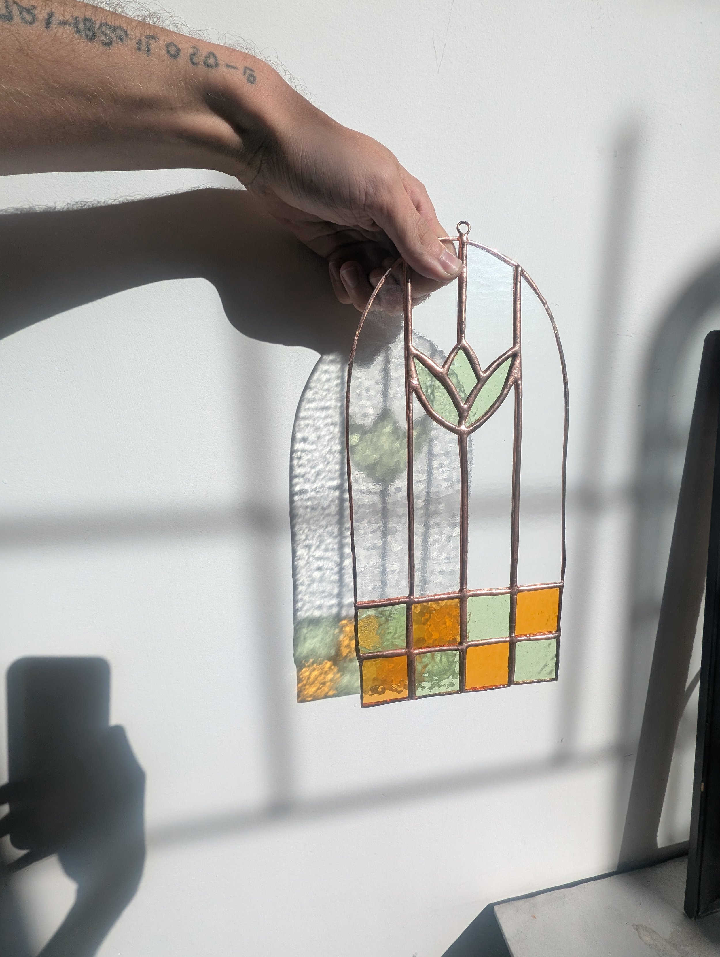 A person's hand holding a stained glass piece with a floral design, casting a colorful shadow on a white wall.