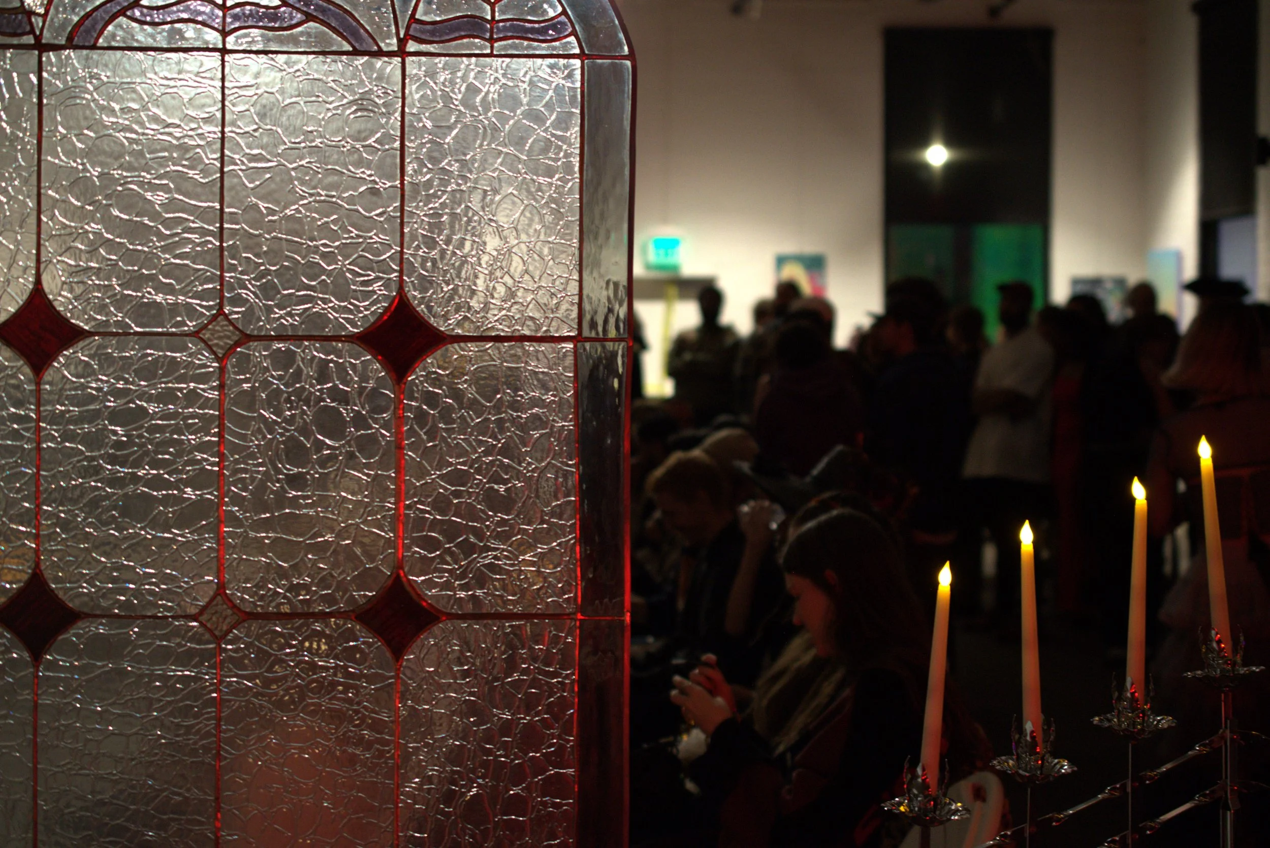 A stained glass panel with red and textured glass in the foreground, and a crowd of people in a dimly lit indoor space in the background, some sitting and some standing, with candles and artwork visible.