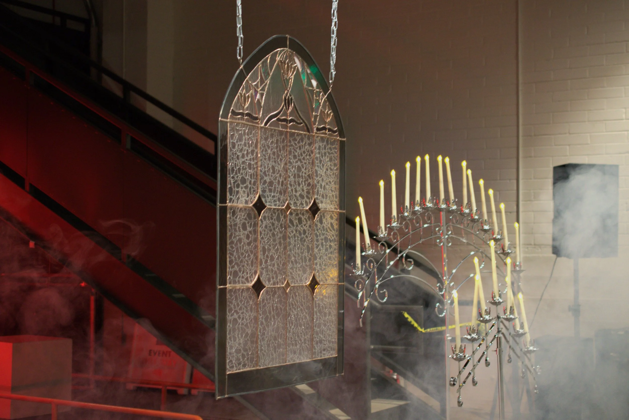 A large decorative stained glass window hanging next to a candelabra with many lit candles, surrounded by smoke or fog, in an indoor setting.