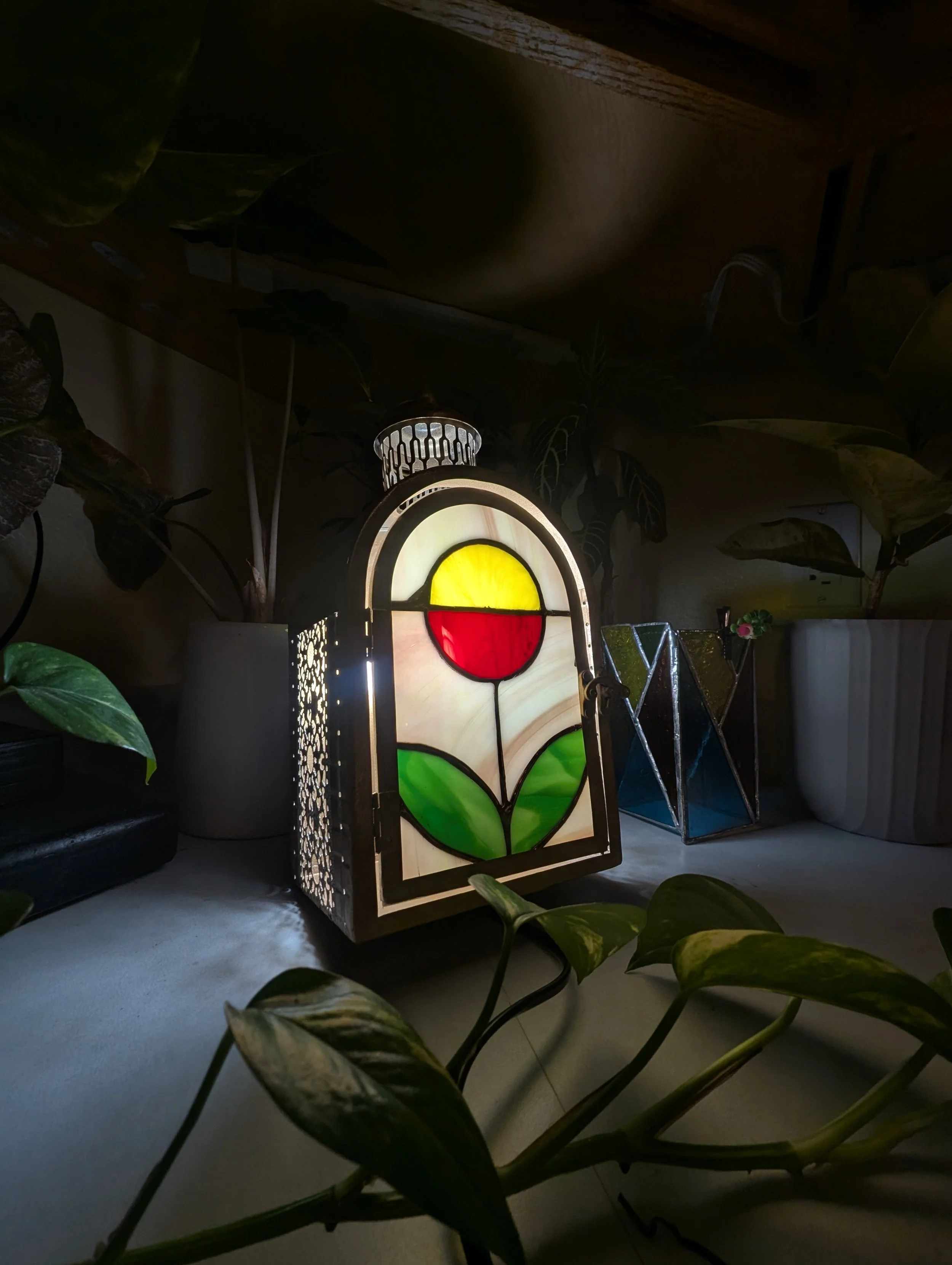 Vase-shaped stained glass lamp with a tulip design, featuring a yellow and red flower with green leaves, placed on a white surface surrounded by houseplants.