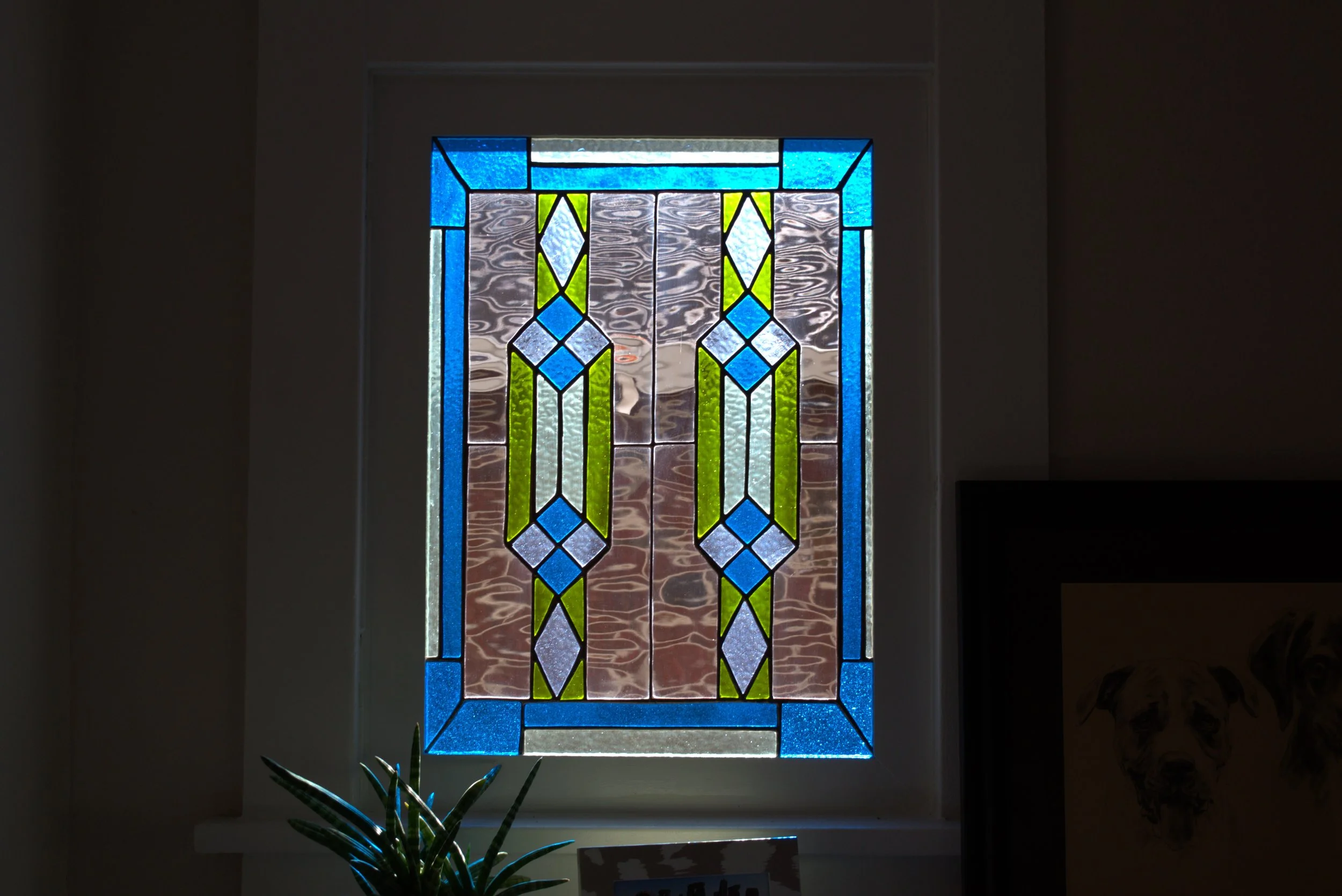 Colorful stained glass window with geometric patterns in blue, green, white, and purple, illuminated by light through the glass.